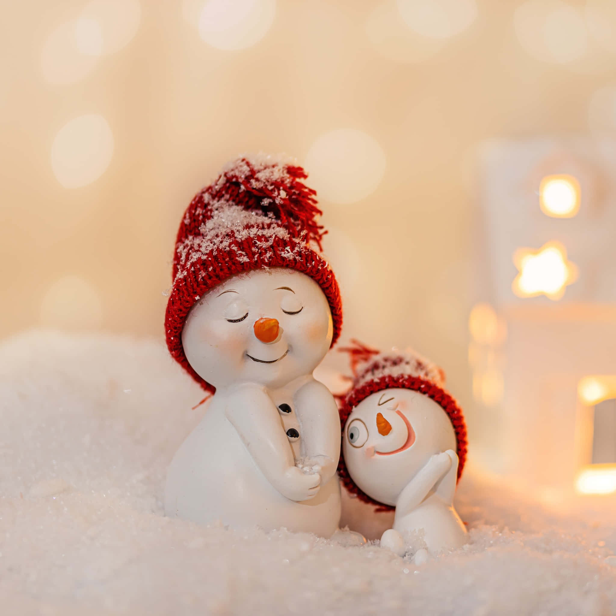 Christmas Eve 桌布 壁紙 — 兩個陶瓷雪人公仔在柔軟的雪中依偎，背景是溫暖發光的燈光和散景效果。