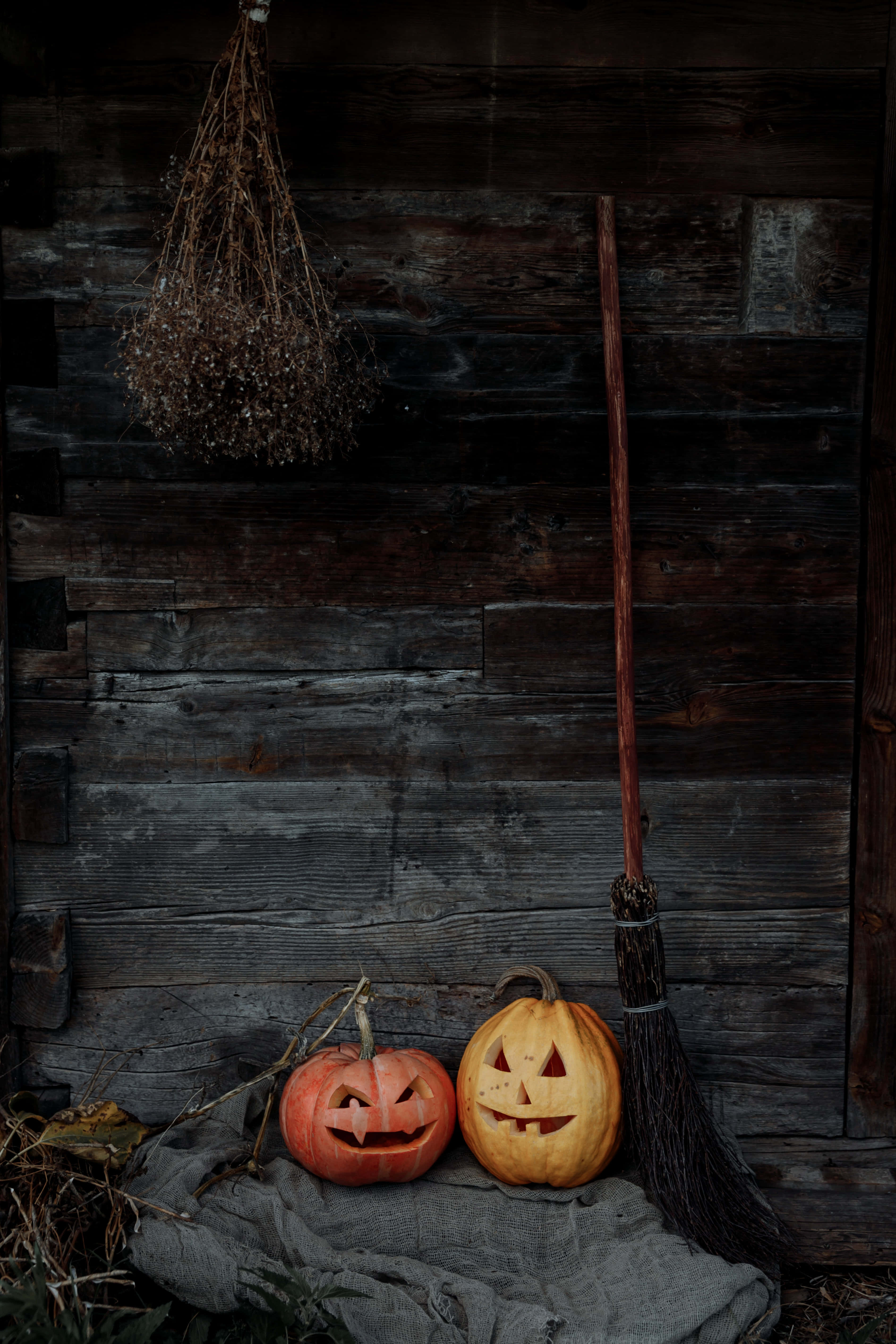 Pumpkin Patch 桌布 壁紙 — 兩個雕刻的南瓜燈和一把掃帚靠在一個風化的木牆上，營造出昏暗的鄉村萬聖節場景。