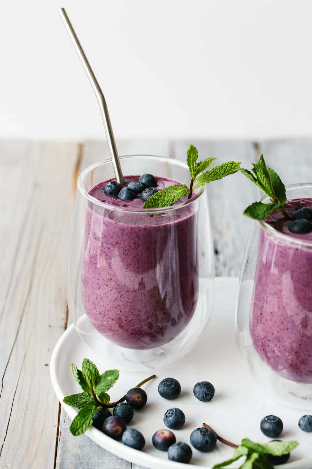 Blueberry Smoothie 桌布 壁紙 — 兩個雙層玻璃杯中的鮮豔紫色藍莓冰沙，飾以新鮮薄荷和整顆藍莓，放在白色盤子上。