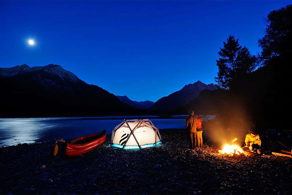 Camping 桌布 壁紙 — 兩個露營者聚集在一個發光的營火旁，旁邊是一個發光的測地線圓頂帳篷，位於月光照耀的湖岸上，遠處是白雪覆蓋的山脈。