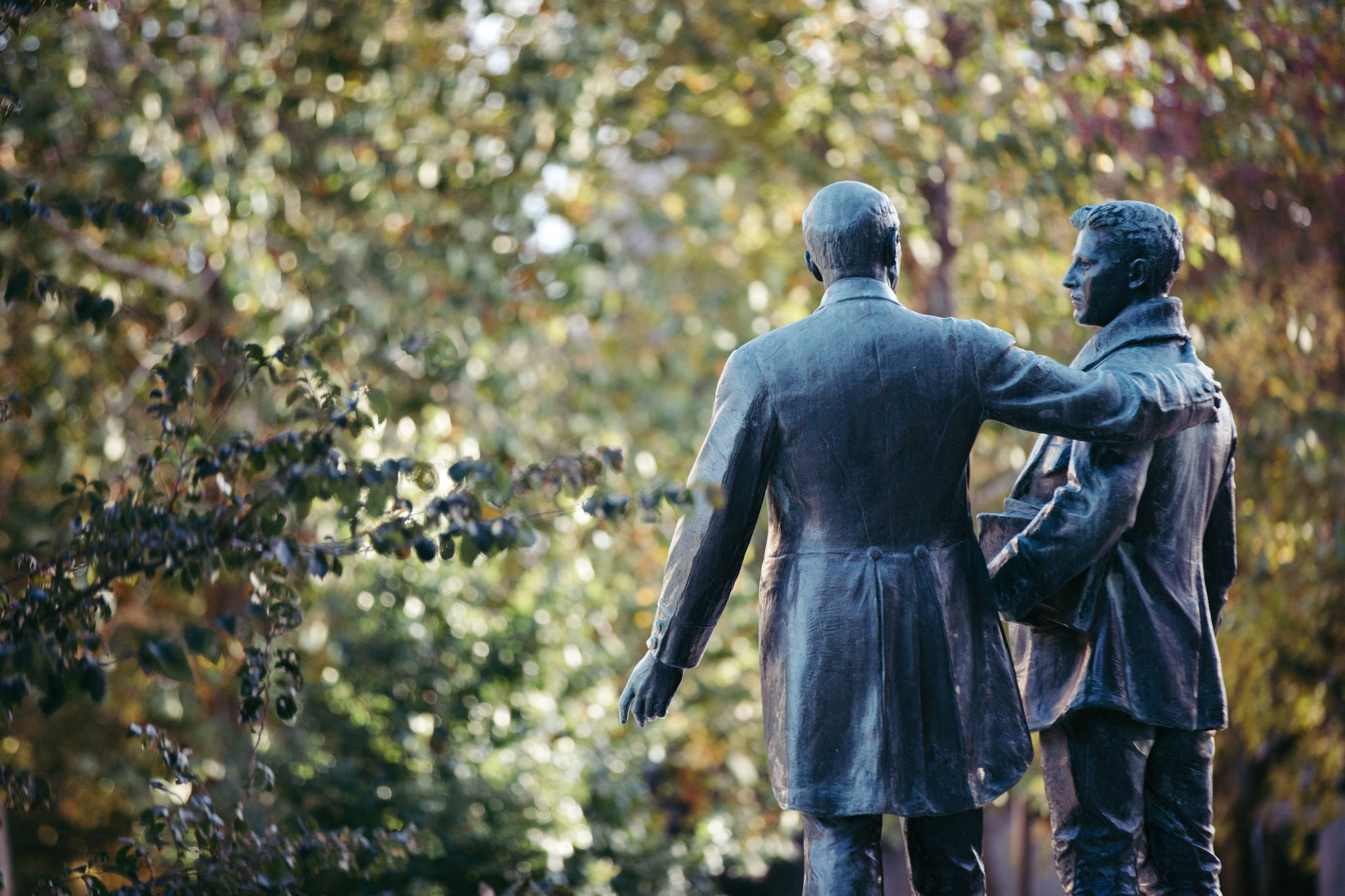 Statue Aesthetic 桌布 壁紙 — 兩個青銅男子雕像並肩走過陽光明媚的秋日森林，樹葉模糊。