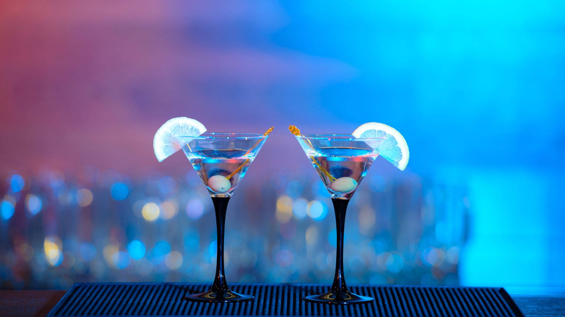 alcohol 桌布 壁紙 — 兩個馬丁尼酒杯，配有橄欖和檸檬角，在充滿活力的藍色和粉紅色霓虹燈下，在酒吧墊上碰杯。
