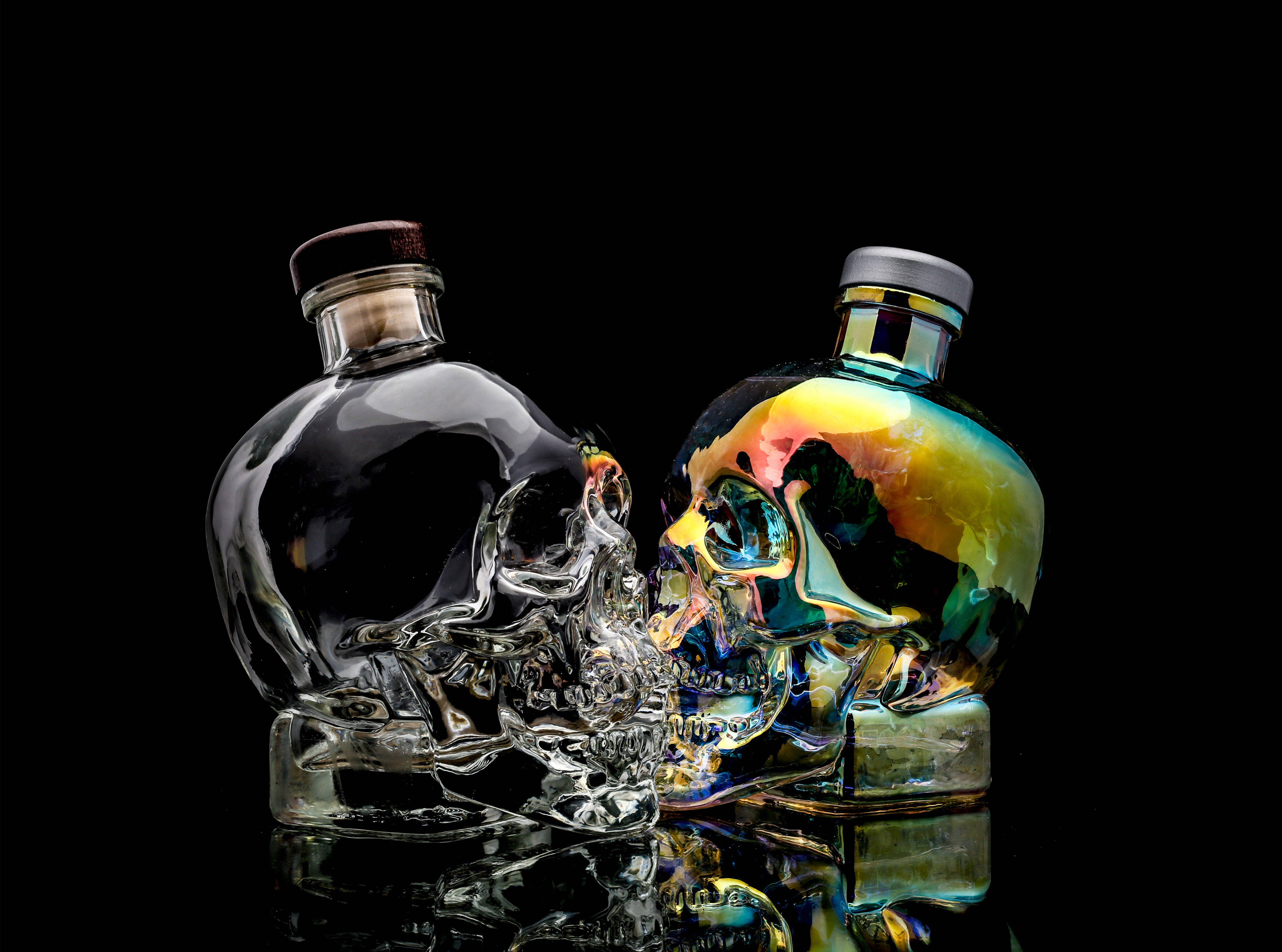 Drink 桌布 壁紙 — 兩個骷髏形狀的酒瓶，一個透明，一個彩虹色，映照在光亮的黑色表面上。