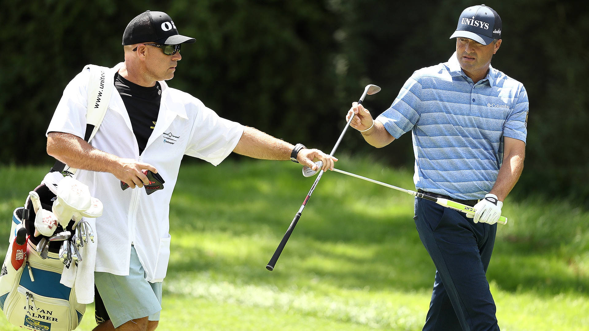 Ryan Palmer 桌布 壁紙 — 兩個高爾夫球手在陽光明媚的球場上，其中一人將球桿遞給另一人，旁邊有一個球童袋。