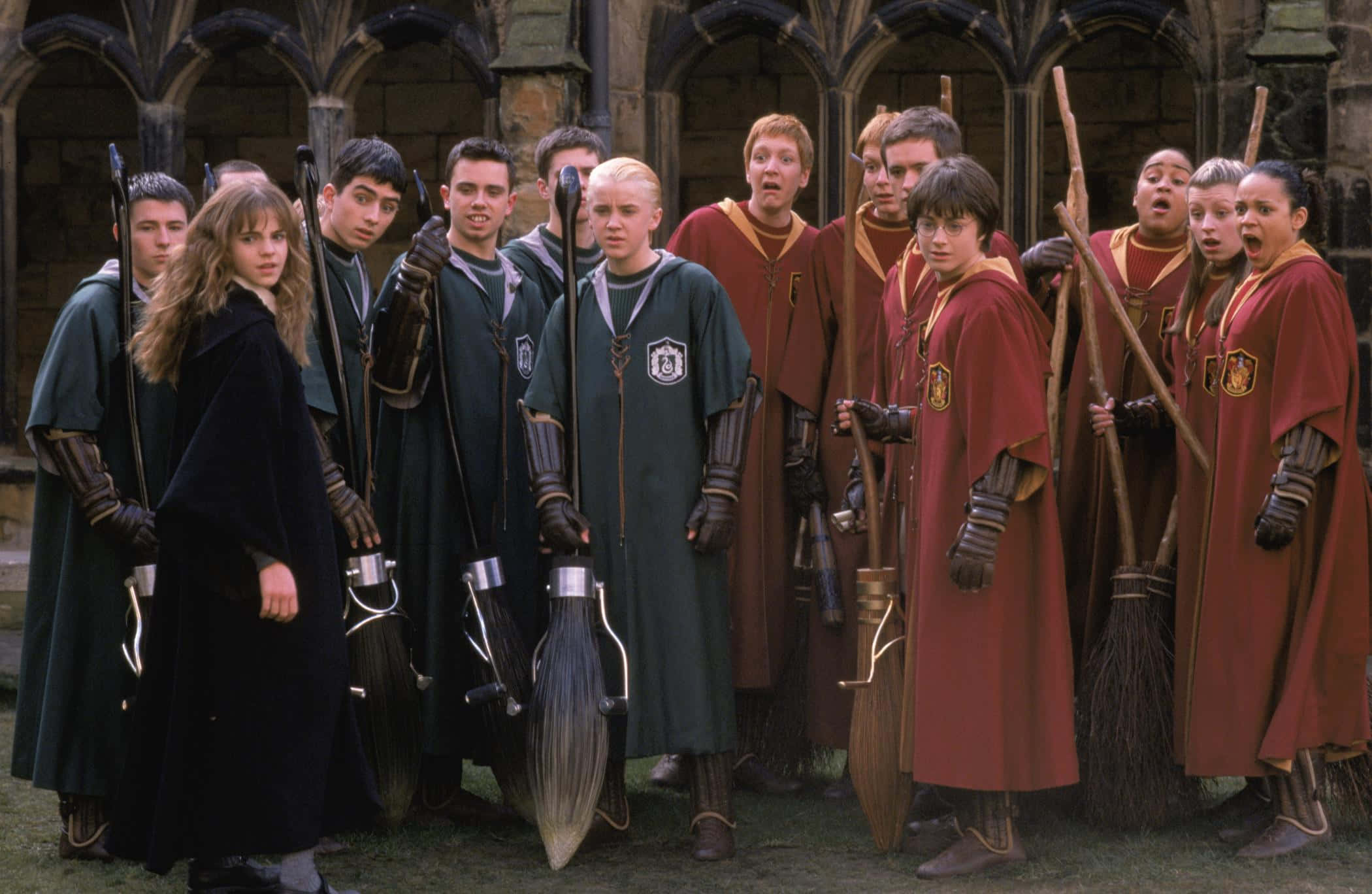 Gryffindor 桌布 壁紙 — 兩個魁地奇隊伍身穿綠色和紅色長袍，手持掃帚站在石頭庭院中，表情緊張而專注。