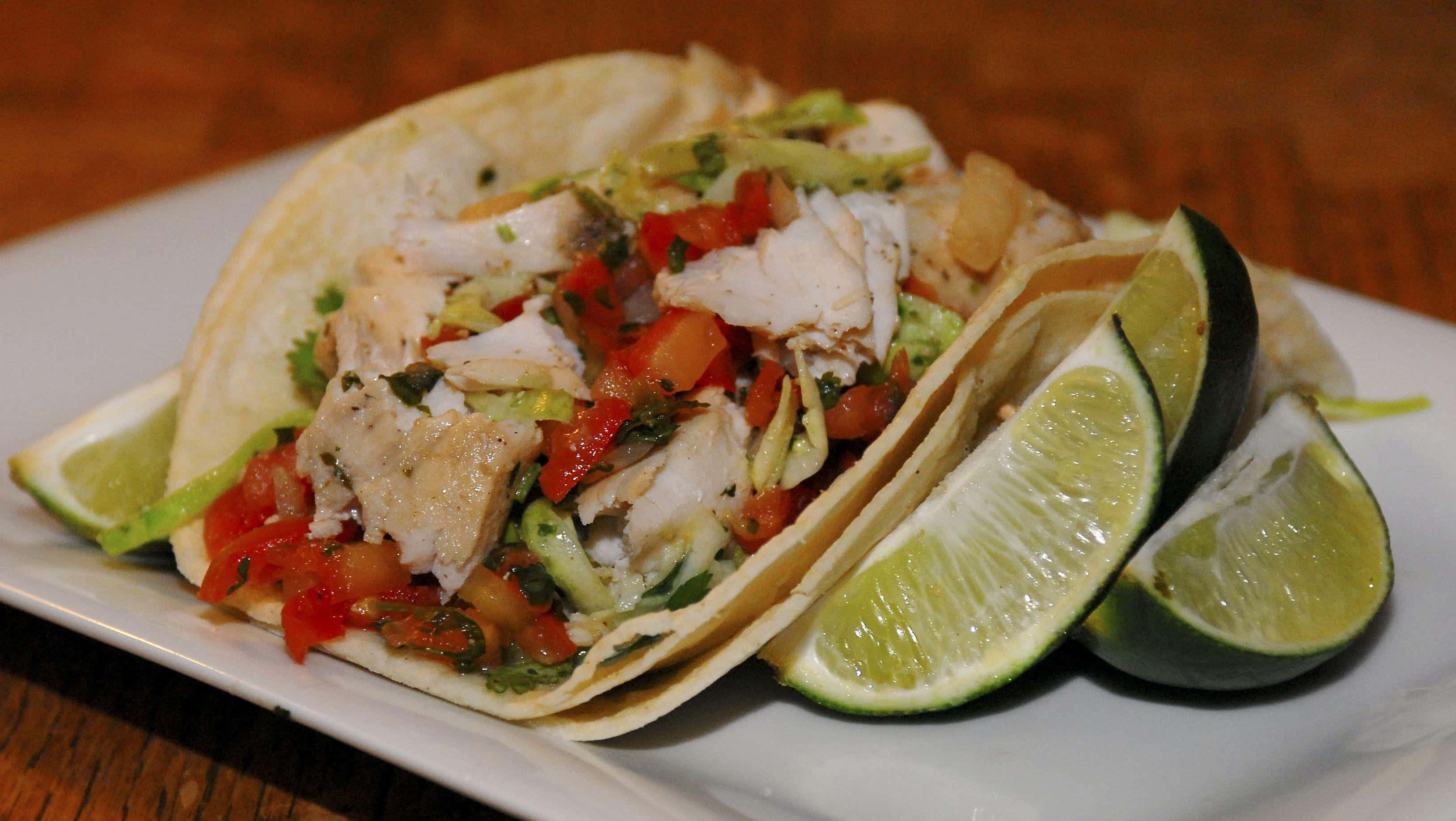 Taco 桌布 壁紙 — 兩個魚肉墨西哥捲，上面放上新鮮的pico de gallo和酪梨，放在白色盤子上，配上萊姆角，置於木桌上。