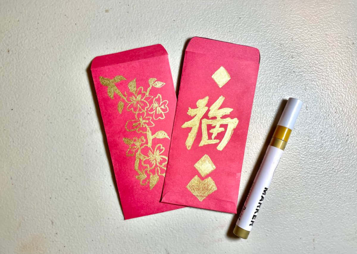 Red Envelope 桌布 壁紙 — 兩個鮮紅色的紅包，上面有金色花卉和福字，旁邊放了一支白色記號筆，在帶有紋理的米白色表面上。