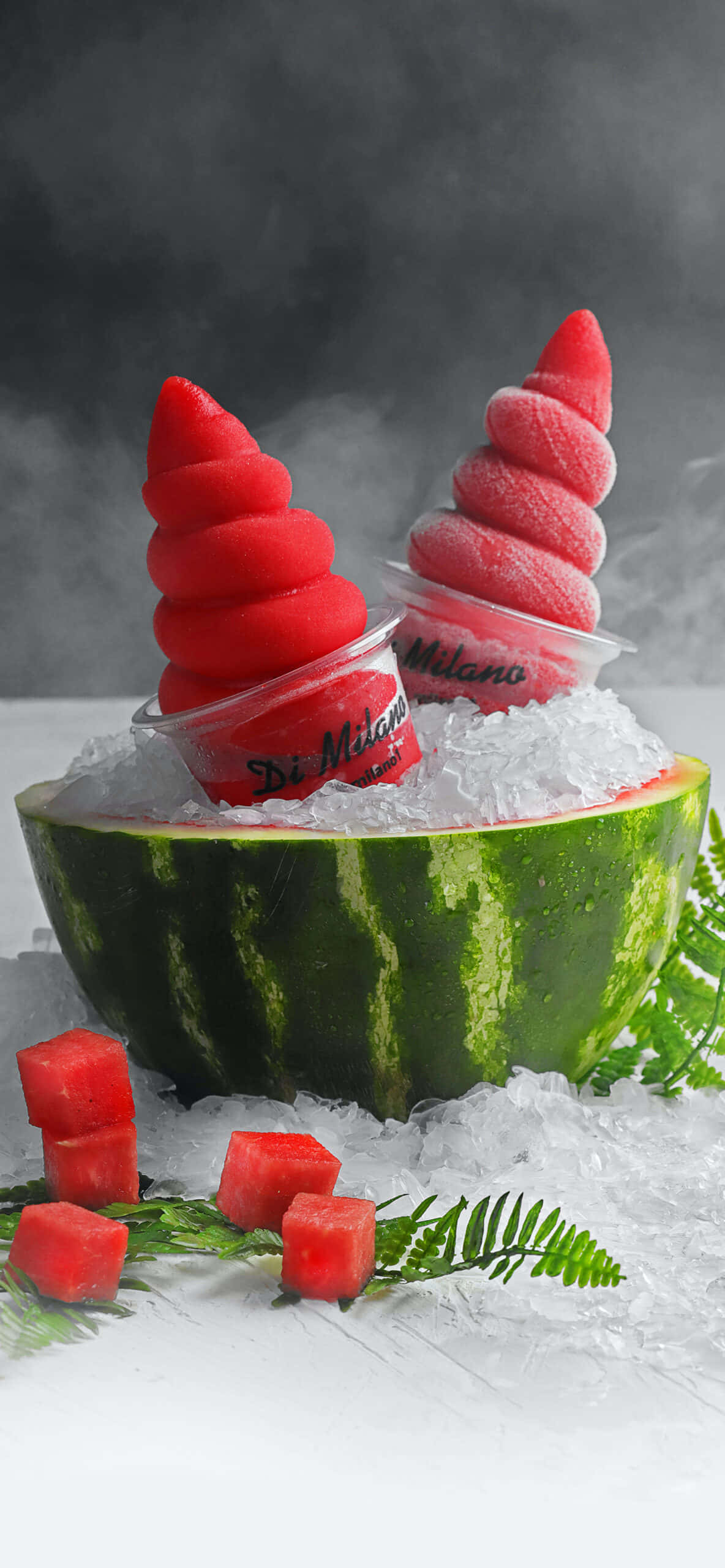 Watermelon iPhone 桌布 壁紙 — 兩個鮮紅色螺旋形冷凍甜品放在挖空的西瓜碗中，碗內裝滿冰塊，並以新鮮西瓜丁和蕨類植物點綴。