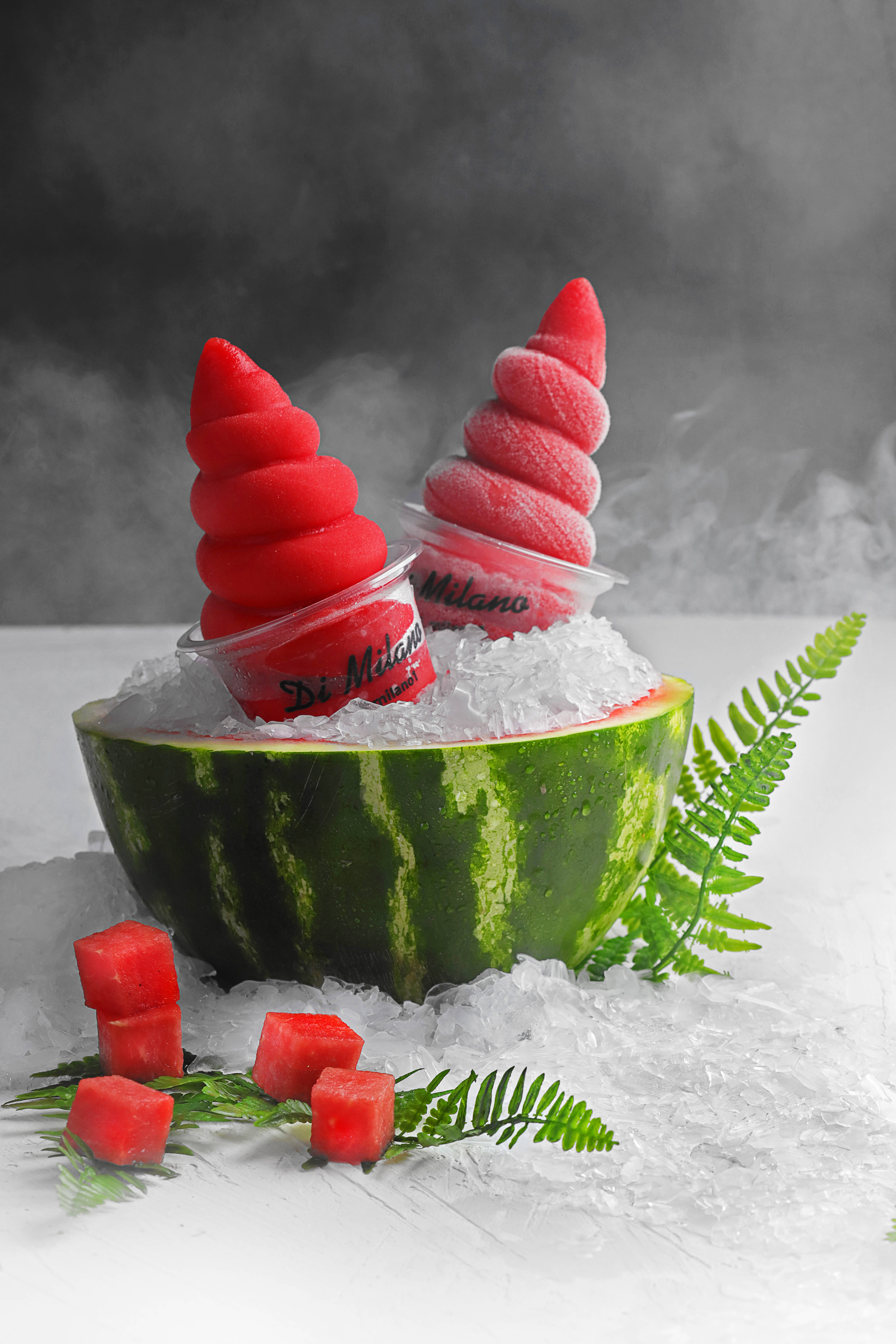 Watermelon 桌布 壁紙 — 兩個鮮紅色螺旋西瓜冰淇淋甜筒放在一個挖空的西瓜碗中，碗內裝滿冰塊，蒸氣在深灰色背景前升起。