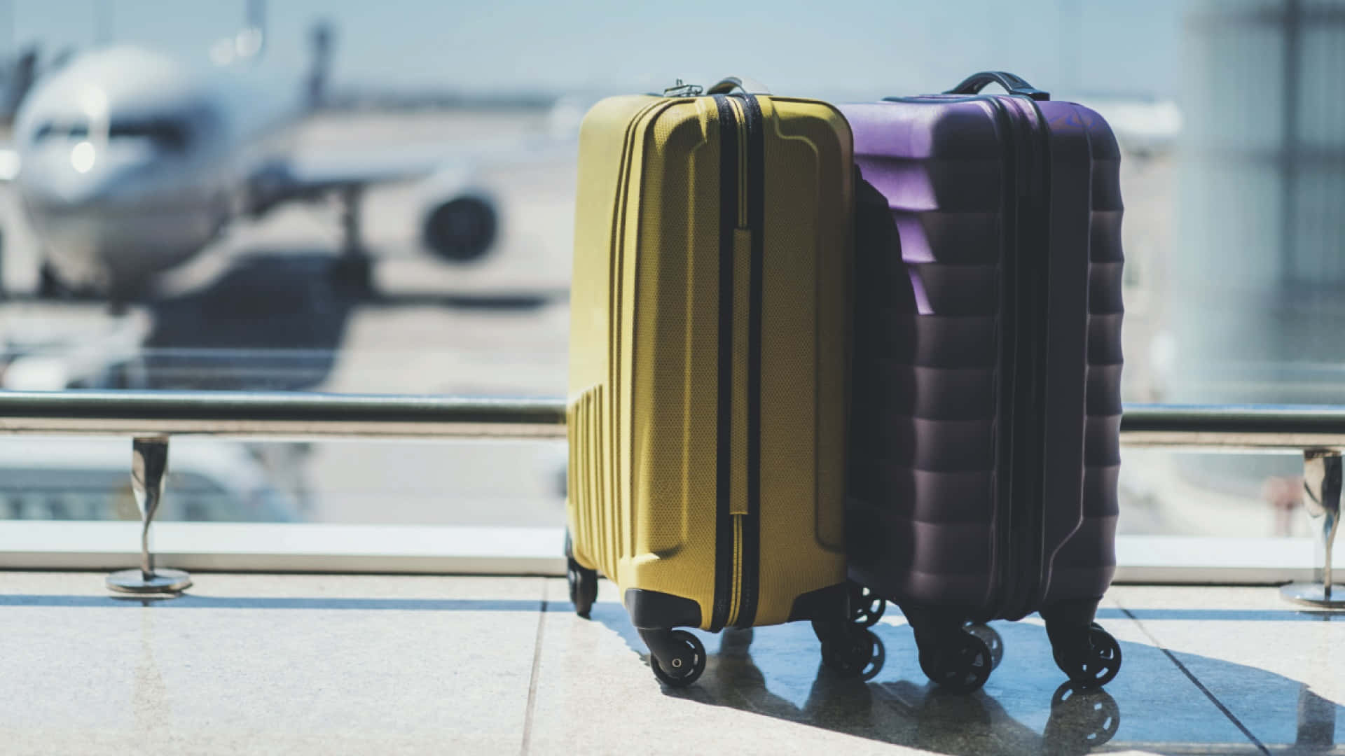 Purple Travel Bag 桌布 壁紙 — 兩個鮮黃色和紫色的行李箱在陽光普照的機場陽台上準備就緒，俯瞰遠處的飛機。