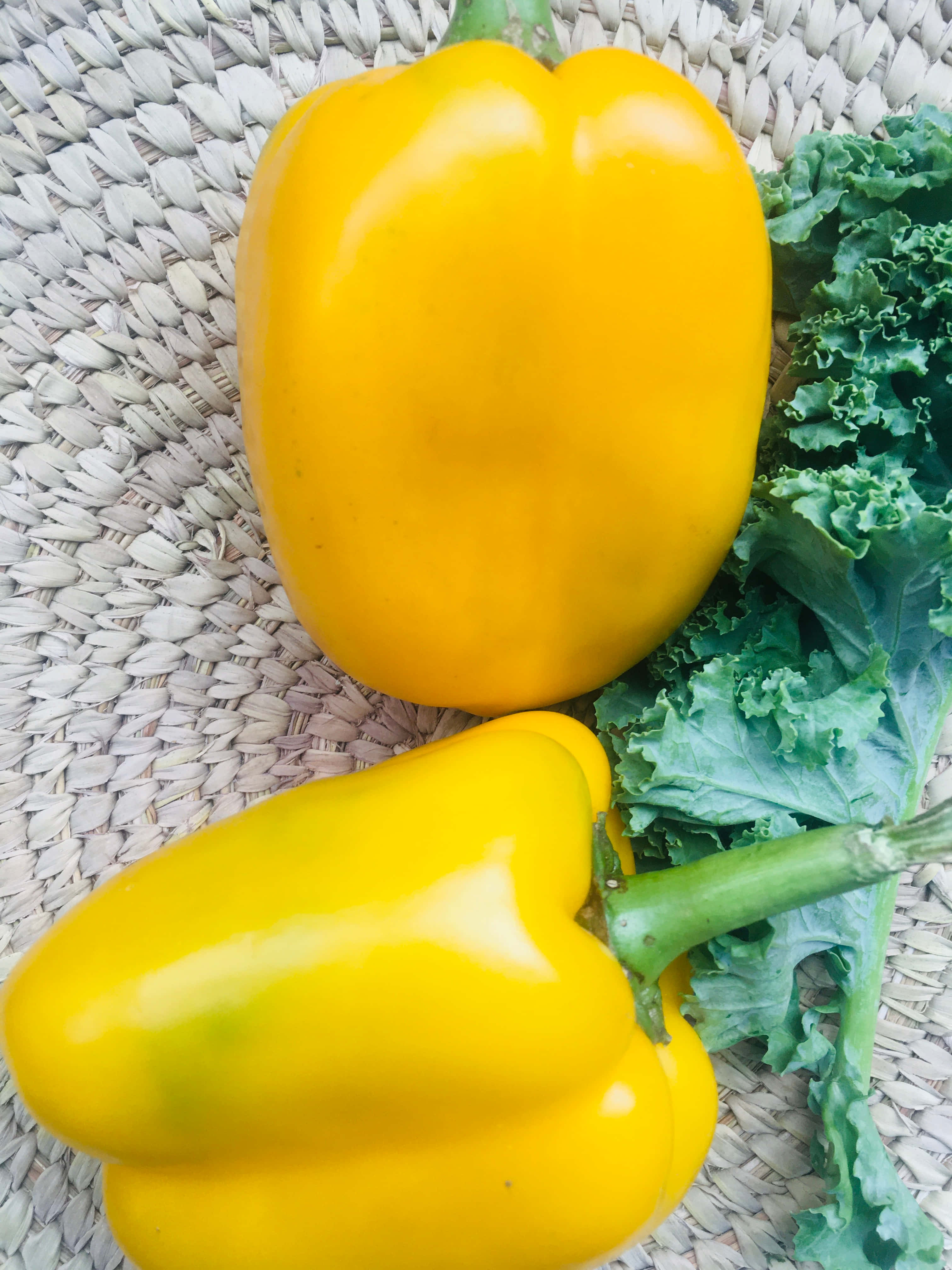 Yellow Bell Pepper 桌布 壁紙 — 兩個鮮黃色甜椒和羽衣甘藍葉放在編織草蓆上，在明亮的自然光下。
