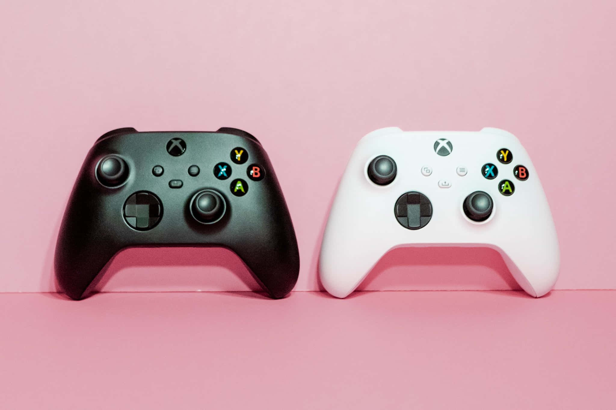 Xbox 桌布 壁紙 — 兩個黑色和白色的Xbox控制器並排放在柔軟的粉紅色背景上。