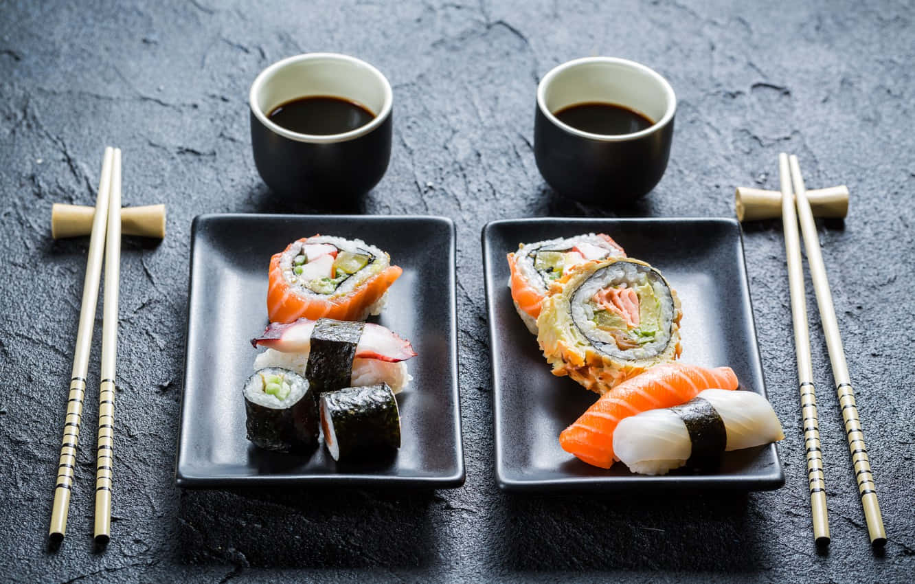 Sushi 桌布 壁紙 — 兩個黑色盤子盛裝各種壽司卷和握壽司，附有醬油杯和筷子，放在深色紋理表面上。