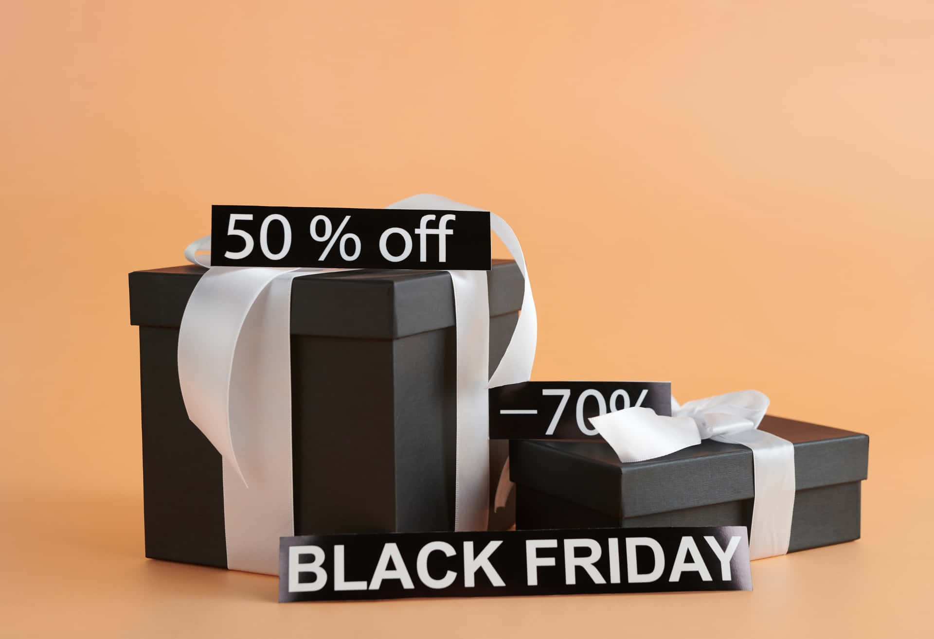 Black Friday Sale Gift Boxes with Discount Tags 壁紙 — 兩個黑色禮物盒，帶白色絲帶，展示折扣標牌，分別為50% off和70% off，背景為溫暖的桃色，並有Black Friday橫幅。
