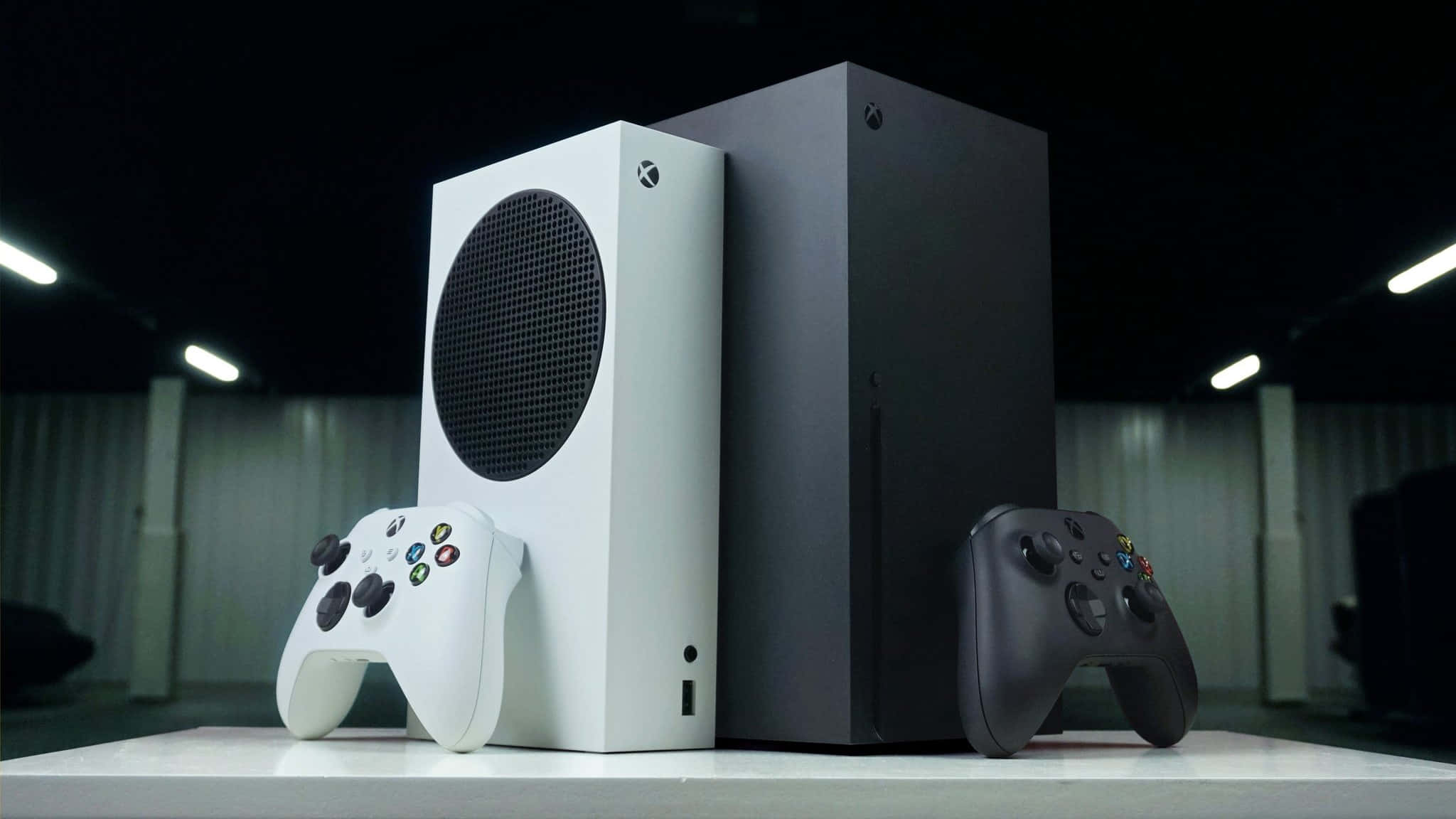 Xbox 桌布 壁紙 — 兩台 Xbox 主機和控制器並排放在白色表面上，上方有明亮的燈光。
