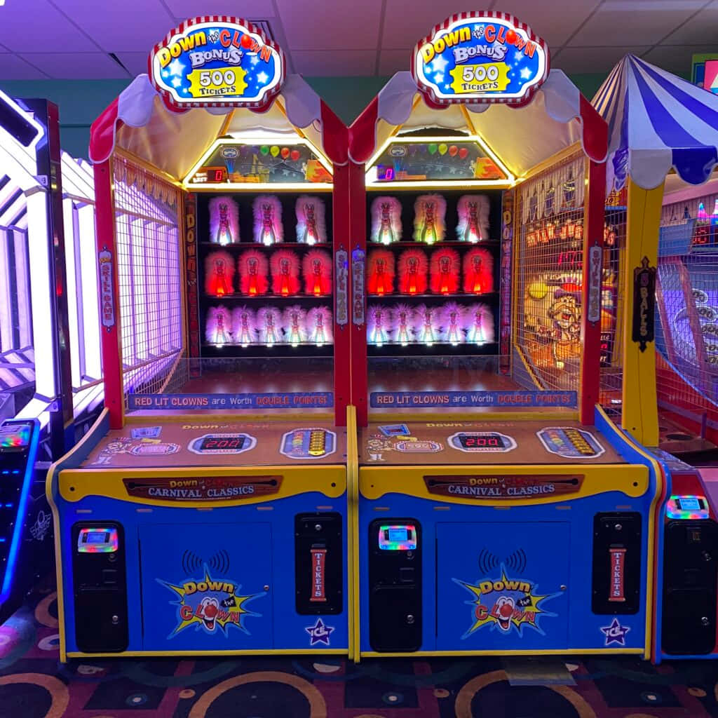 Arcade Games 桌布 壁紙 — 兩台燈光明亮的嘉年華遊戲機，內部有發光的紅色和紫色小丑公仔，位於色彩繽紛的遊戲機中心。