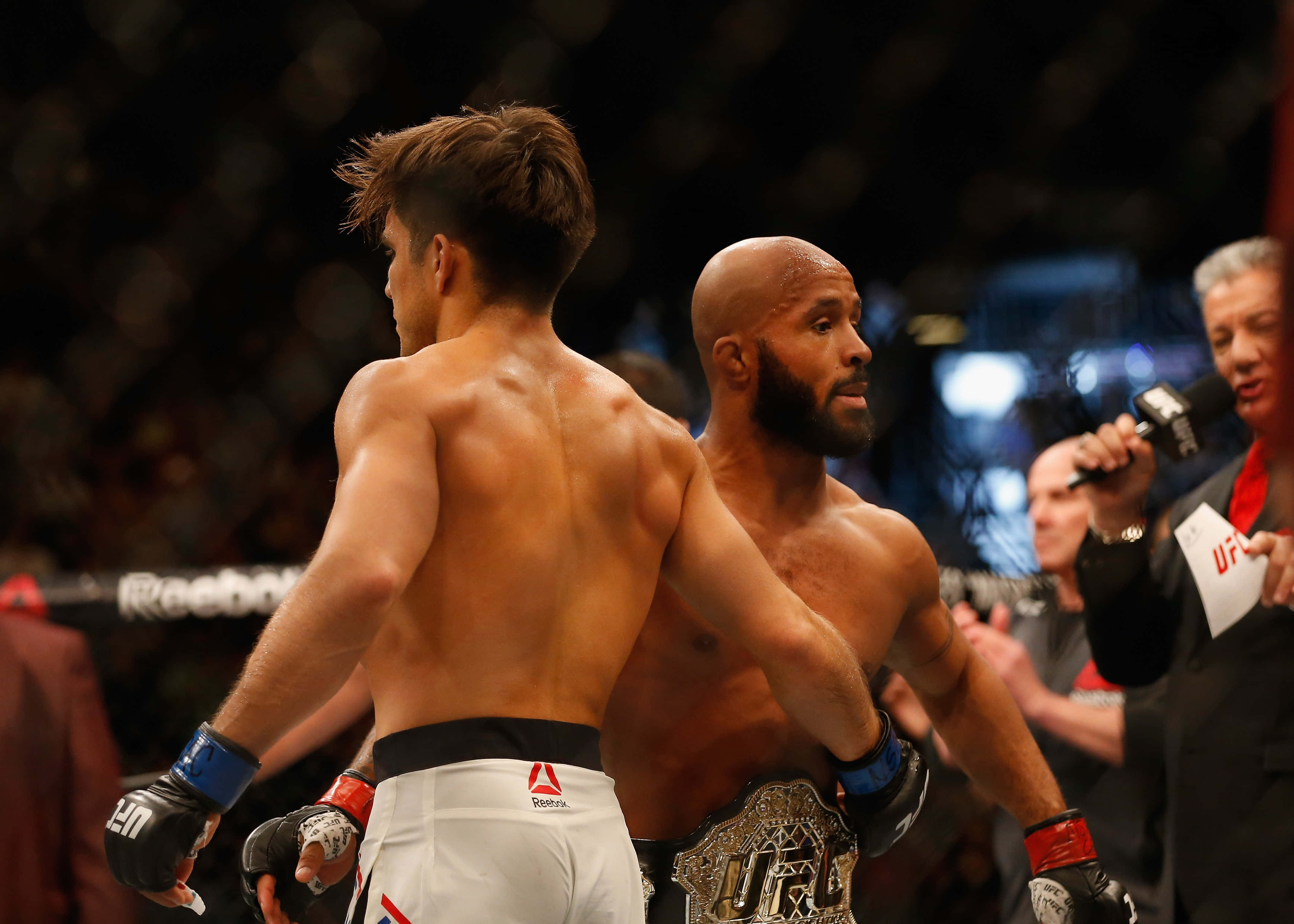 UFC 桌布 壁紙 — 兩名MMA拳手在籠中對峙，其中一人手持冠軍腰帶，背景是明亮的場館燈光。