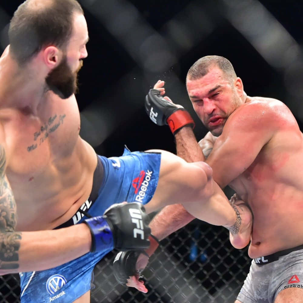 MMA 桌布 壁紙 — 兩名MMA拳手在籠中激烈交鋒，一人使出高踢，另一人阻擋，汗水在昏暗的場館燈光下飛濺。