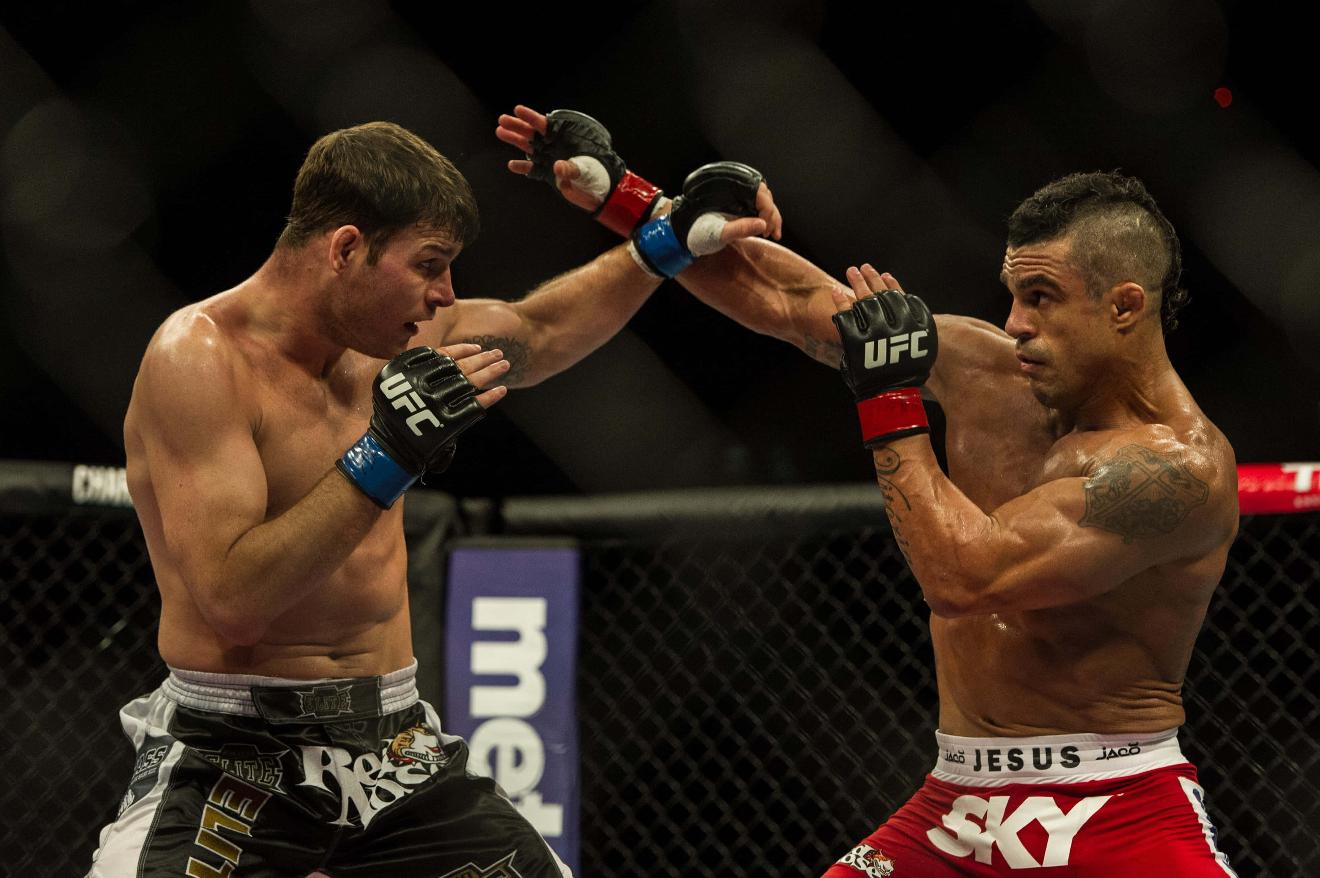 UFC 桌布 壁紙 — 兩名MMA拳手在鐵籠中激烈交鋒，汗水飛濺，其中一人在明亮的場館燈光下向對方臉部擊出強力一拳。