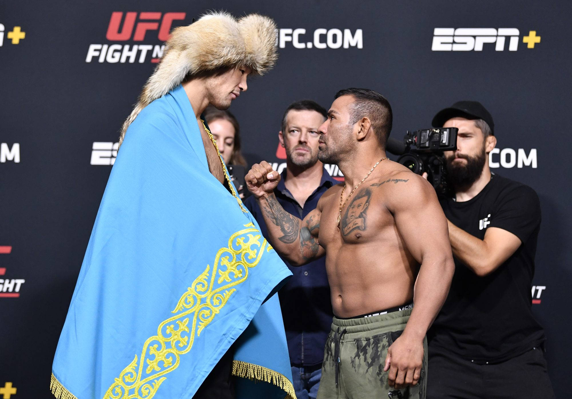 UFC 桌布 壁紙 — 兩名UFC拳手在稱重時對峙，一人披著藍色裝飾旗幟，戴著毛帽，另一人赤裸上身，有刺青和戴金鏈。