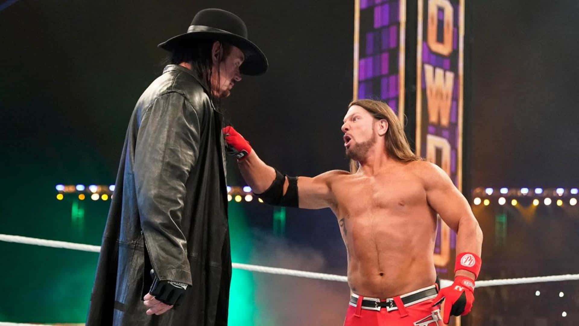 WWE 桌布 壁紙 — 兩名WWE摔角手在擂台上對峙，一人身穿黑色風衣和帽子，另一人赤裸上身，戴紅色手套和穿紅色褲子。