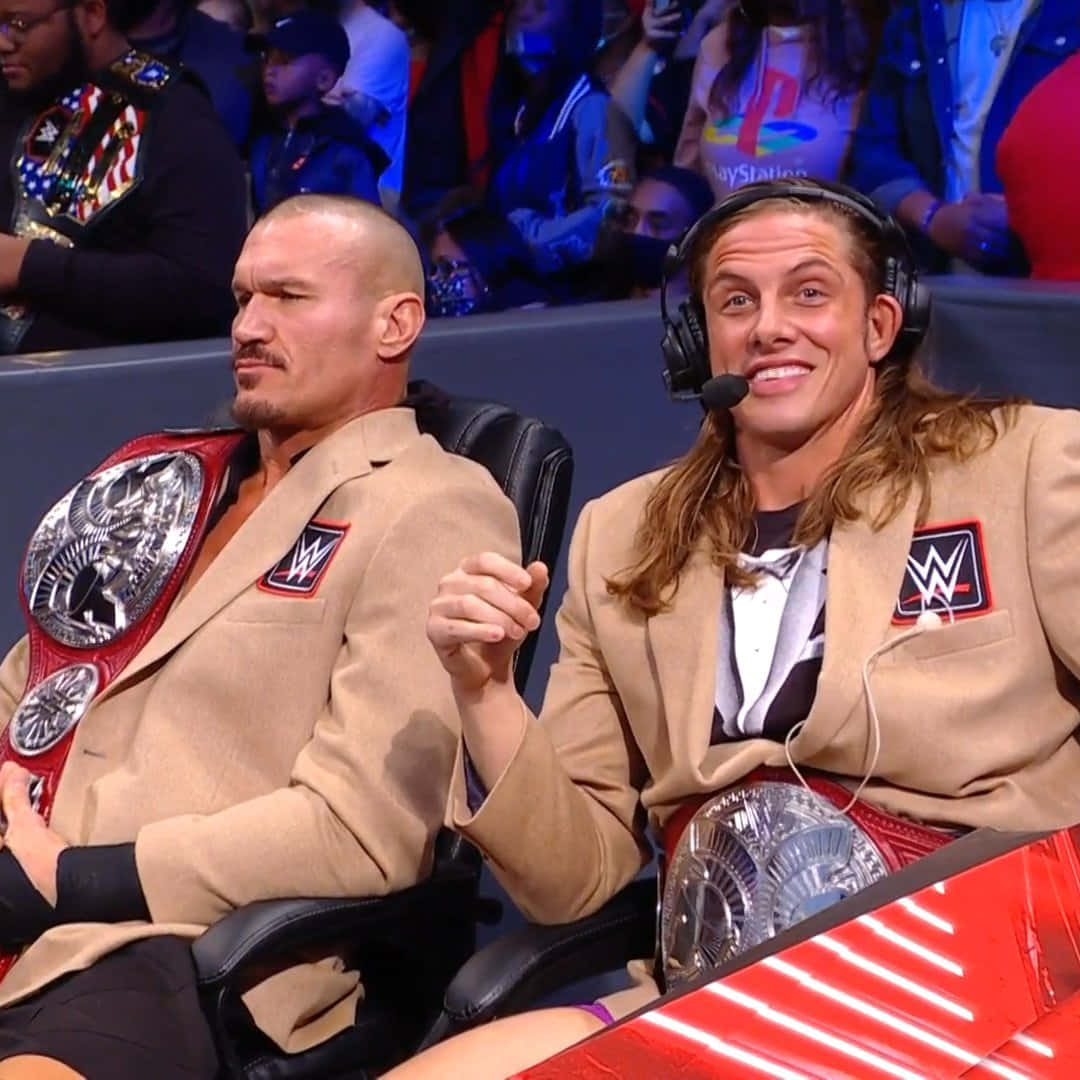 WWE 桌布 壁紙 — 兩名WWE摔角手身穿棕褐色西裝，並排而坐，手持冠軍腰帶，身後有觀眾的現場活動。