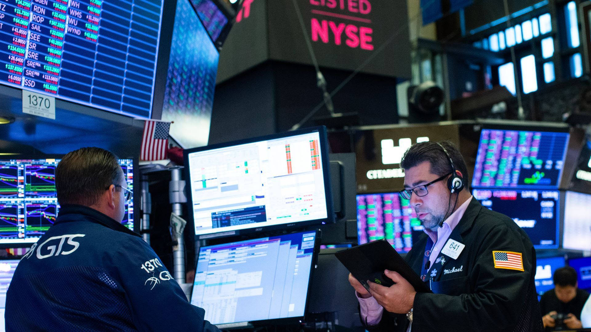 Market 桌布 壁紙 — 兩名交易員在紐約證券交易所交易大廳，在明亮的人造燈光下，專注地盯著顯示股票行情和圖表的發光螢幕。