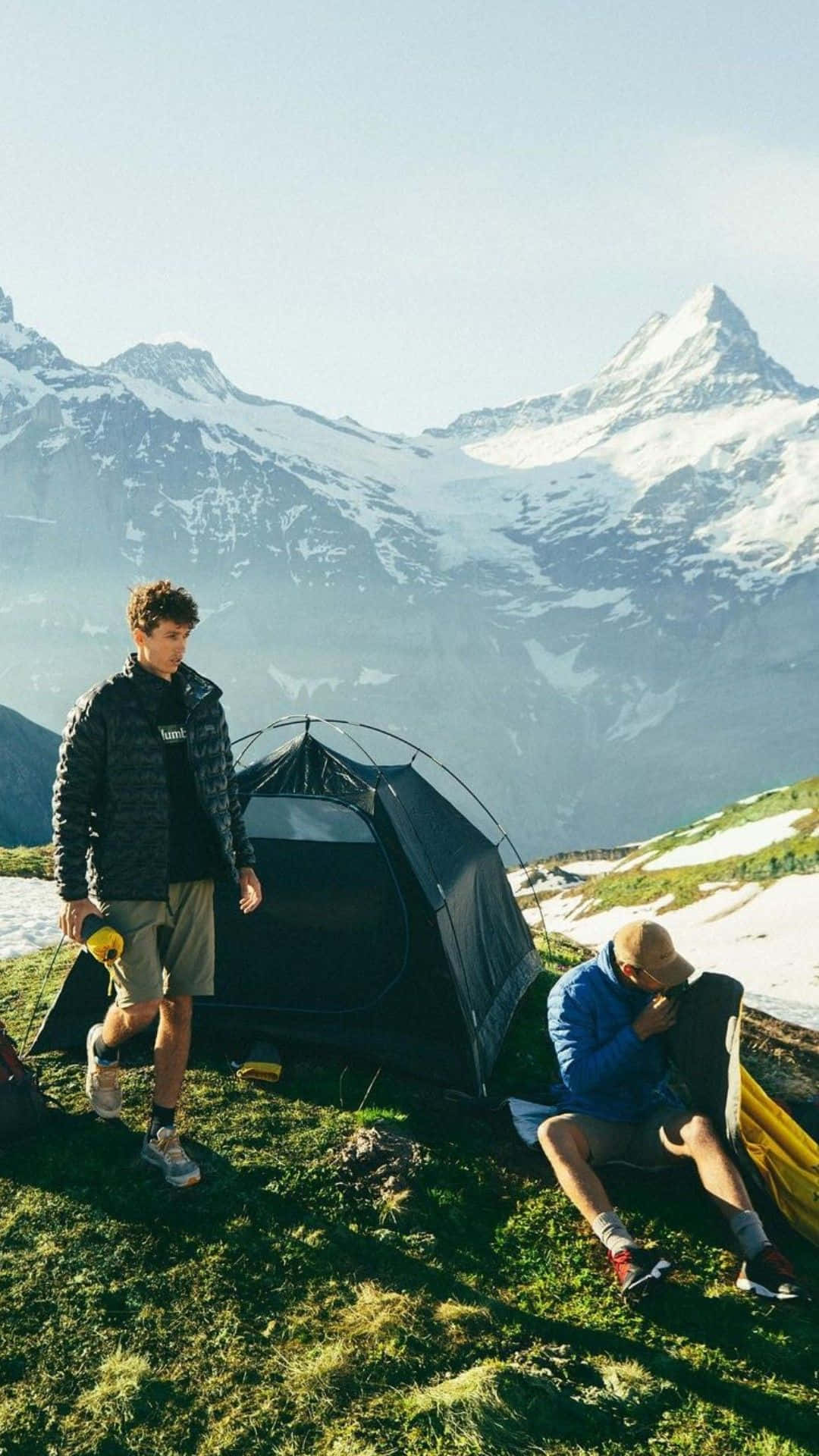 Minimalist Camping 桌布 壁紙 — 兩名健行者在陽光照耀的草地上，靠近一個黑色帳篷休息，背景是白雪覆蓋的山峰高聳入雲。