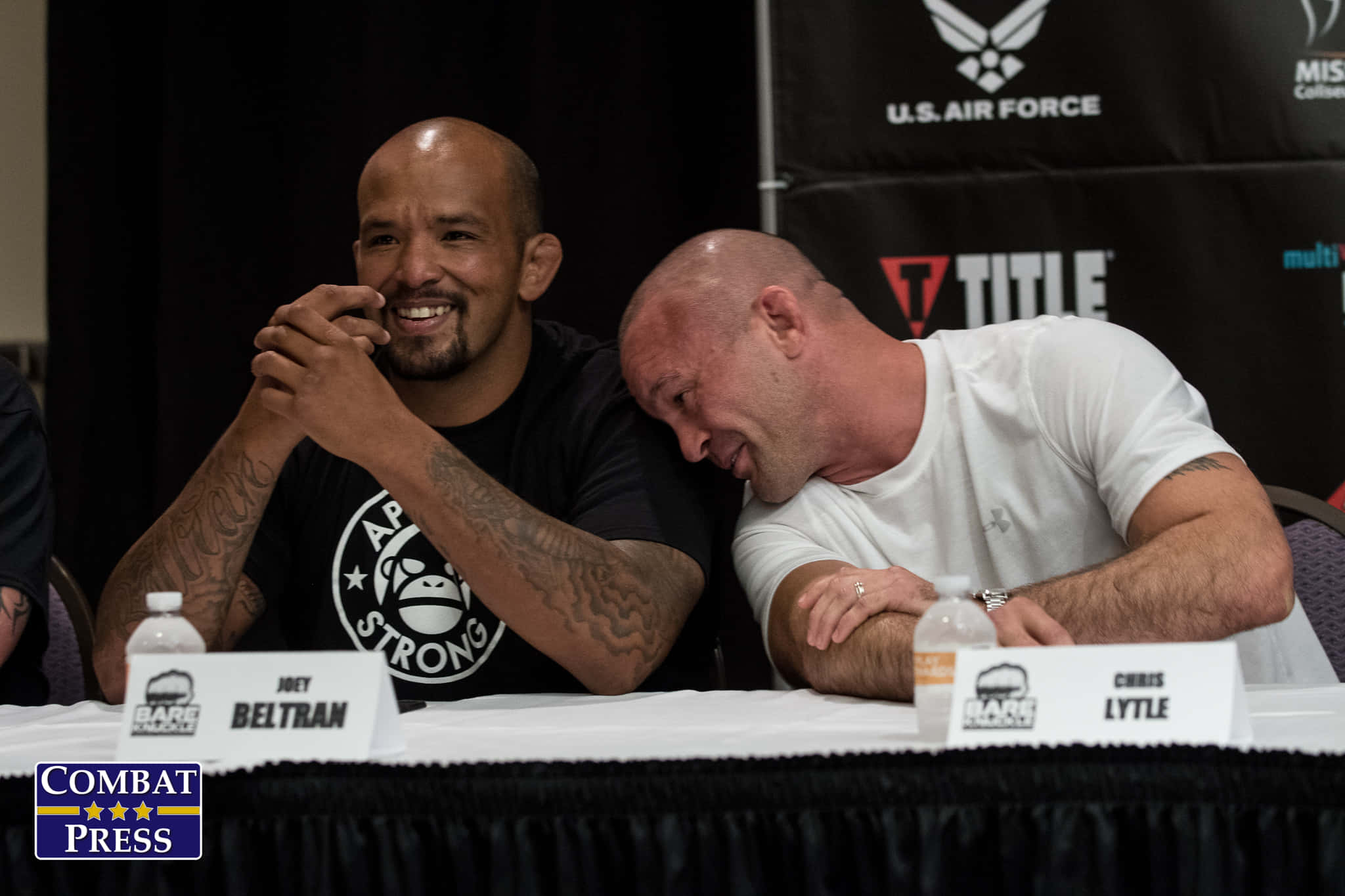 Boxing 桌布 壁紙 — 兩名光頭紋身男子在記者會桌上一起大笑，桌上有名牌和水瓶。