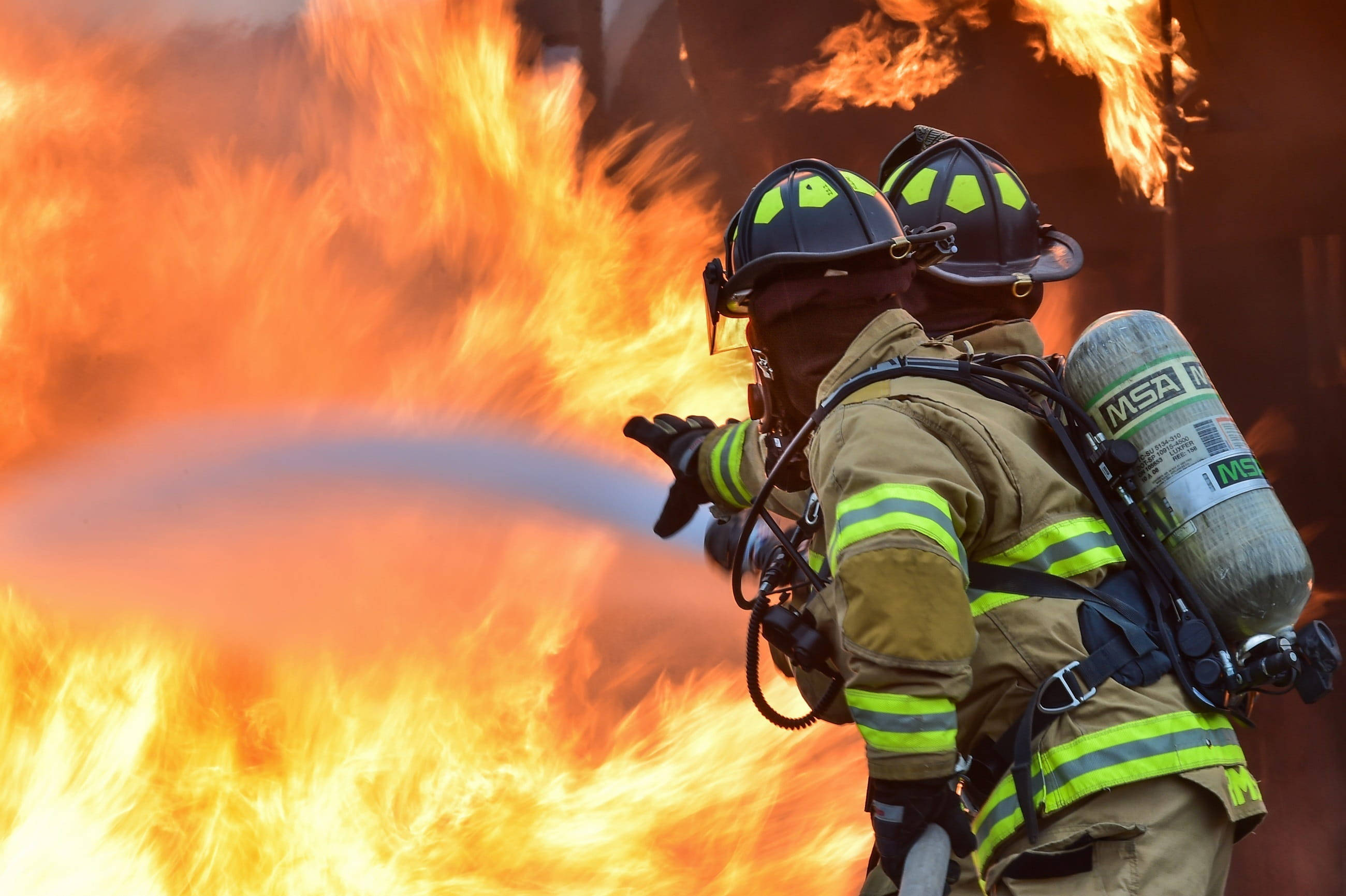 Firefighter 桌布 壁紙 — 兩名全副裝備的消防員在強烈的橙色和黃色火焰背景下，用強勁的水流對抗咆哮的火焰。