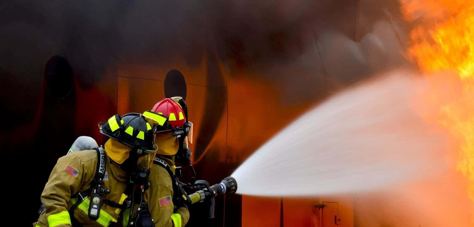 Fire Department 桌布 壁紙 — 兩名全副裝備的消防員在橙色火焰和濃煙的背景下，用強勁的水流對抗熊熊烈火。