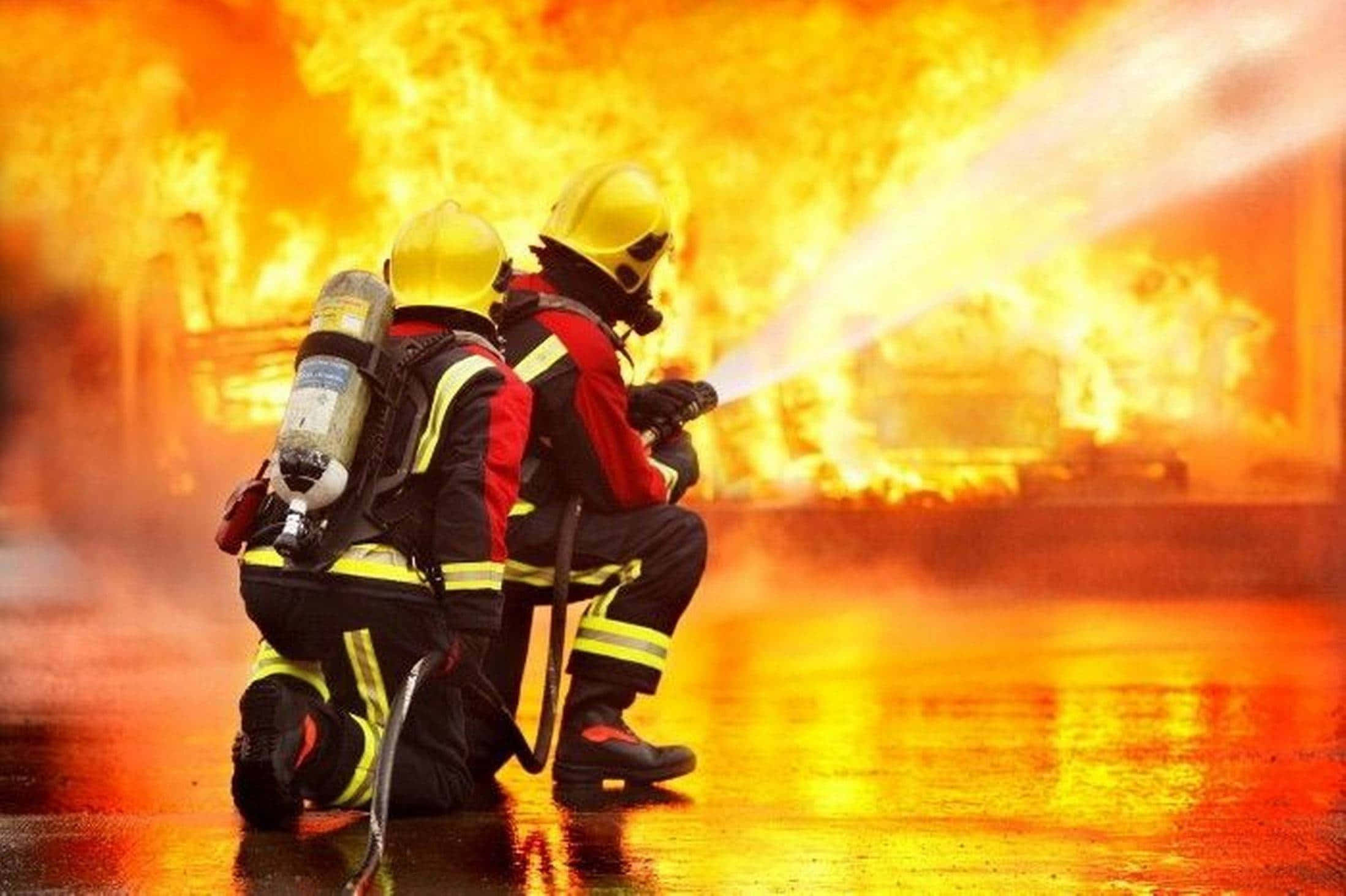 Fire Department 桌布 壁紙 — 兩名全副裝備的消防員跪下，向猛烈的地獄之火噴水，濕漉漉的路面反射出火熱的橙色。