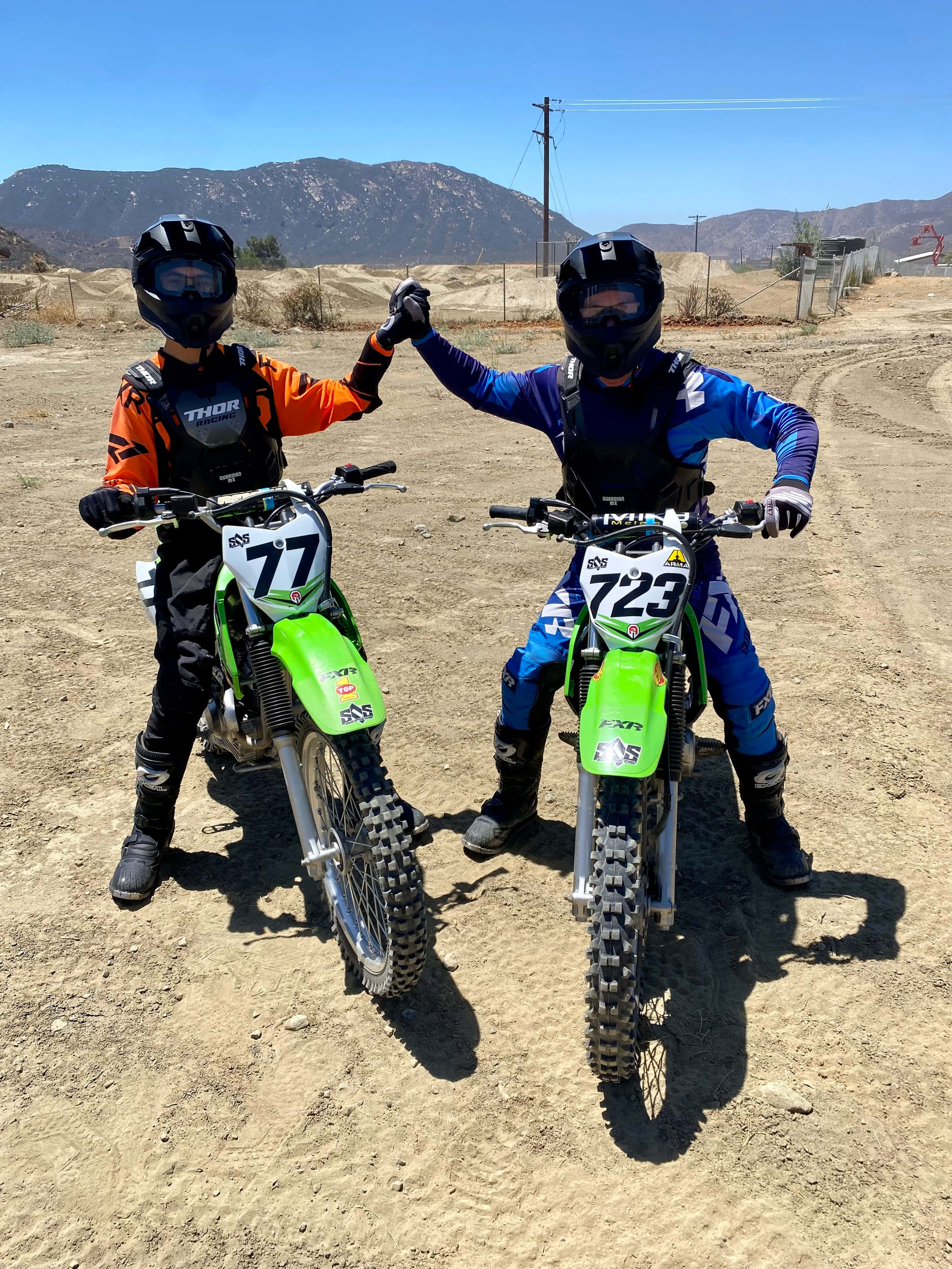 Dirtbike 桌布 壁紙 — 兩名全副裝備的越野摩托車車手在塵土飛揚的賽道上擊掌，背景是山脈和清澈的藍天。