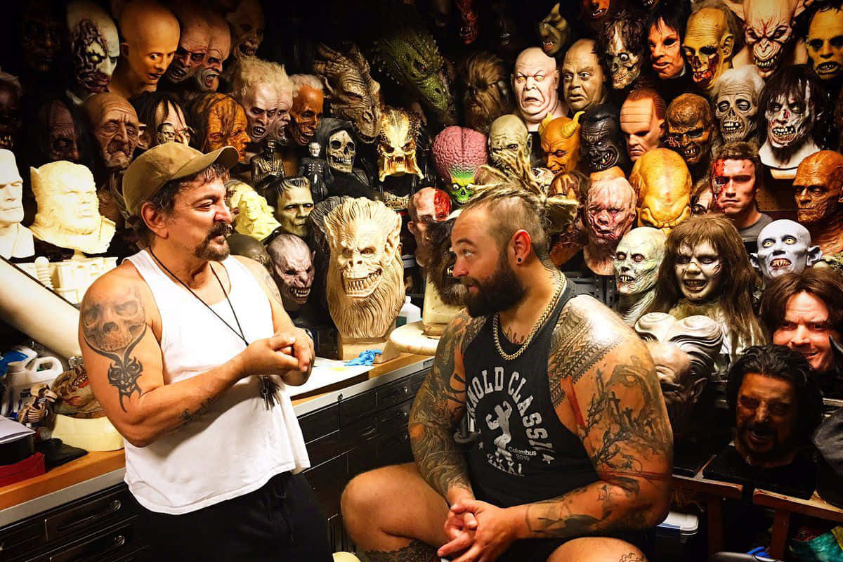 WWE 桌布 壁紙 — 兩名刺青男子在一個雜亂的工作室中交談，周圍環繞著怪誕的恐怖面具。