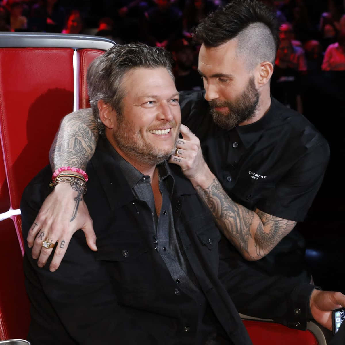 Adam Levine 桌布 壁紙 — 兩名刺青男子在紅色椅子上分享溫馨時刻，身後是模糊的觀眾。