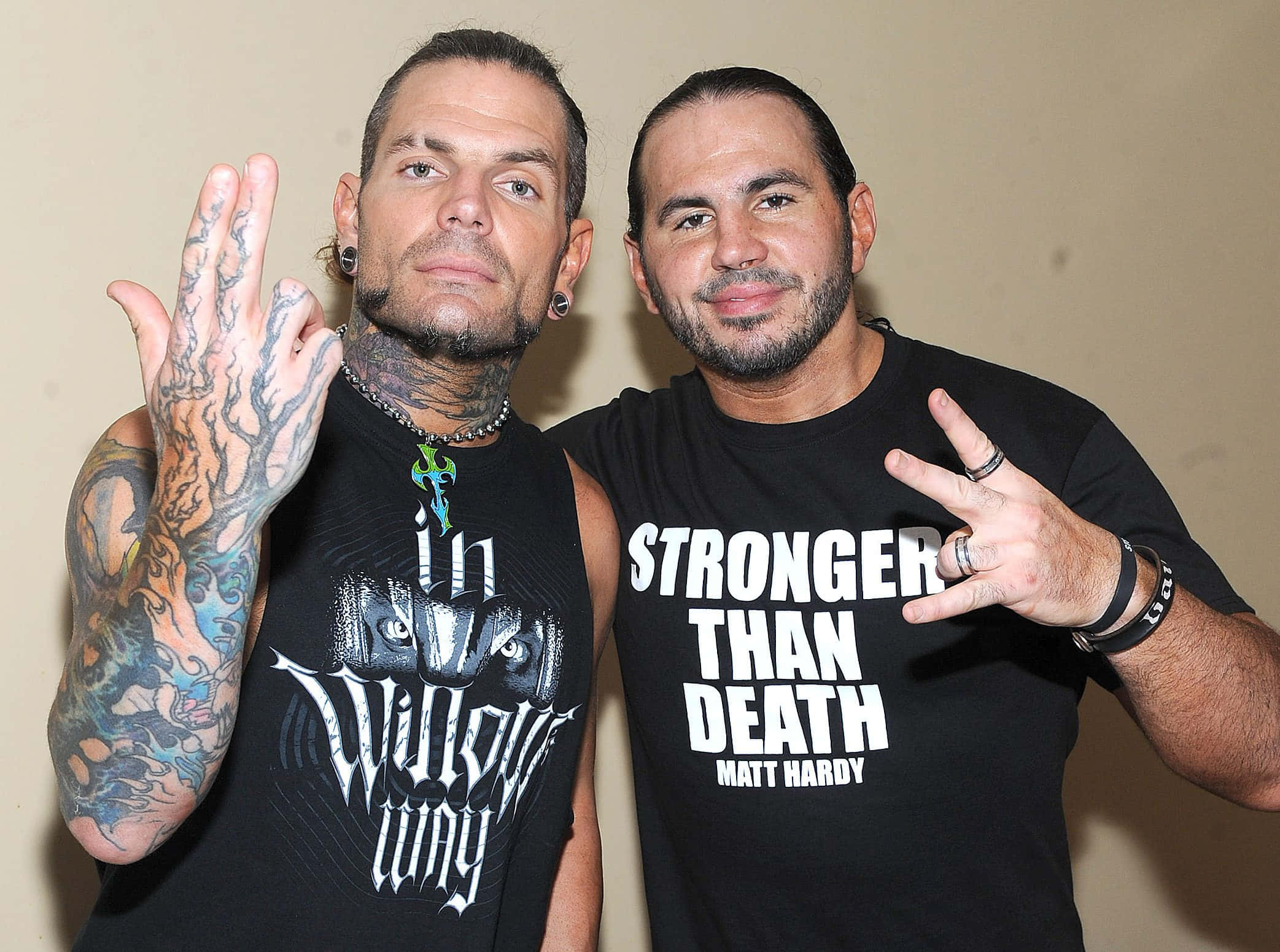 WWE 桌布 壁紙 — 兩名刺青男子自信地穿著黑色T恤擺姿勢，其中一件印有「Stronger Than Death」字樣，兩人均在純色米色牆前比出和平手勢。