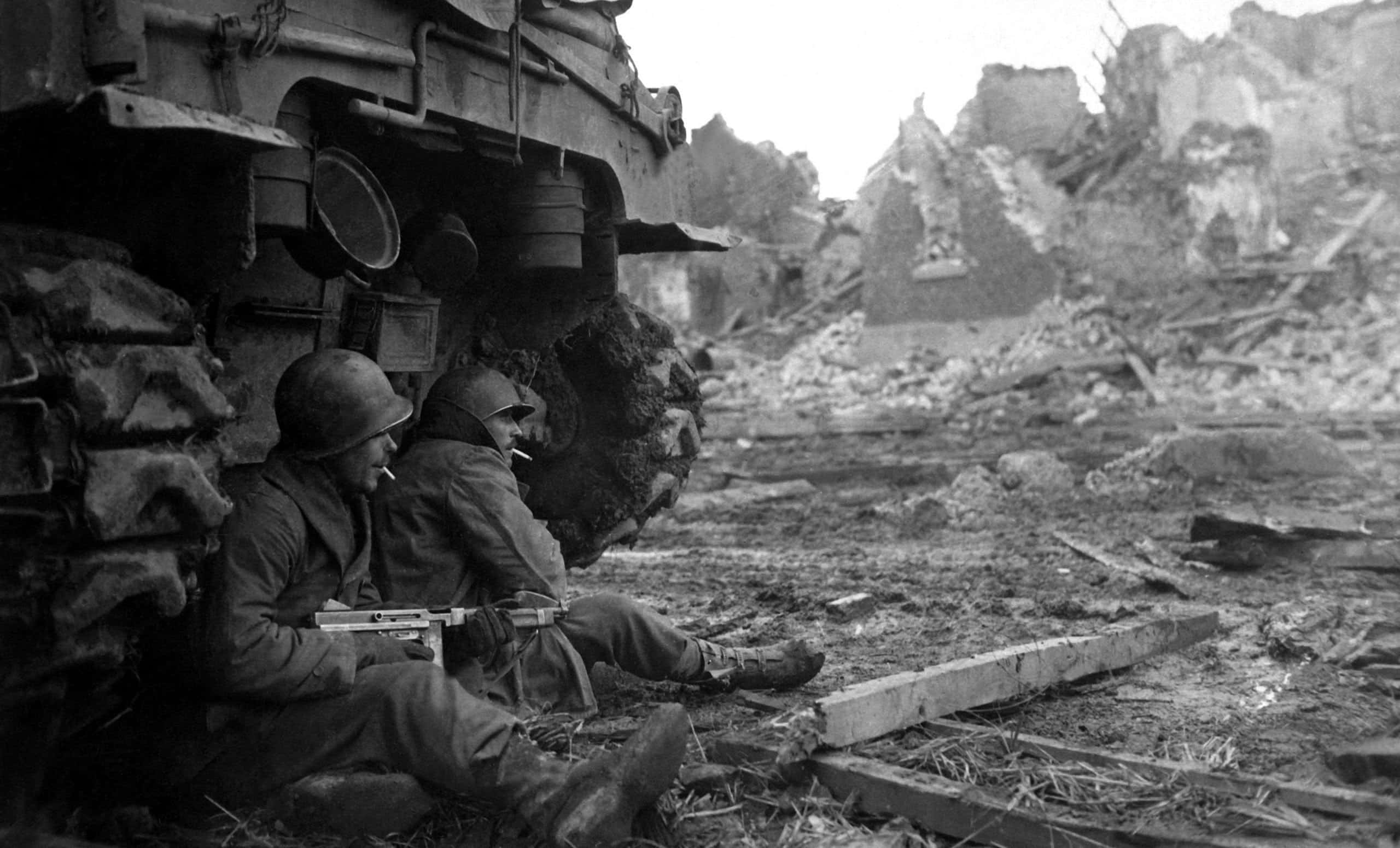 World War 2 桌布 壁紙 — 兩名士兵在戰火摧殘的城市景觀中，在瓦礫和毀壞的建築物旁，於坦克車旁休息。
