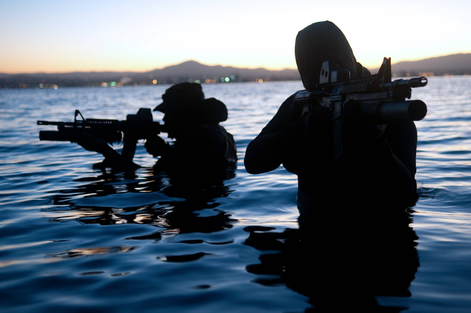 Silhouette 桌布 壁紙 — 兩名士兵在黃昏時分以剪影形式涉過平靜的水面，手持步槍，遠處的城市燈光在地平線上閃爍。