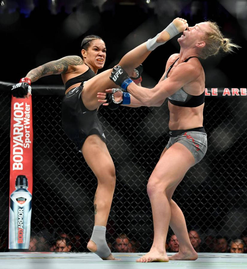 UFC 桌布 壁紙 — 兩名女性MMA拳手在激烈的鐵籠賽中交鋒，其中一人使出高踢，另一人則進行阻擋。