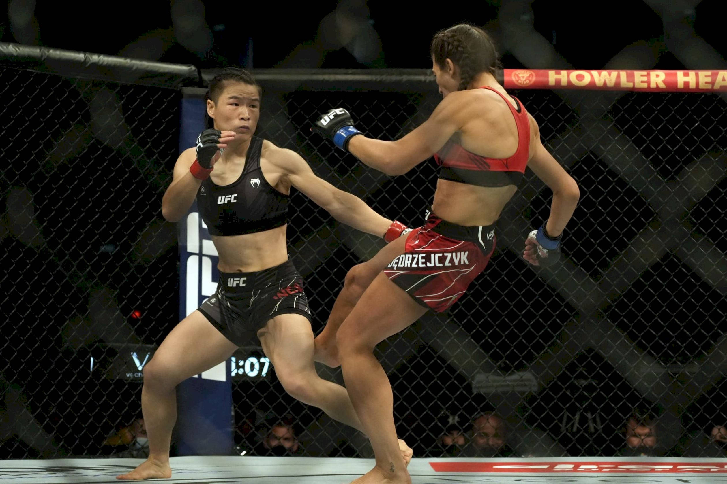 MMA 桌布 壁紙 — 兩名女性MMA拳手在激烈的鐵籠賽中交鋒，一人使出高踢，另一人防守。
