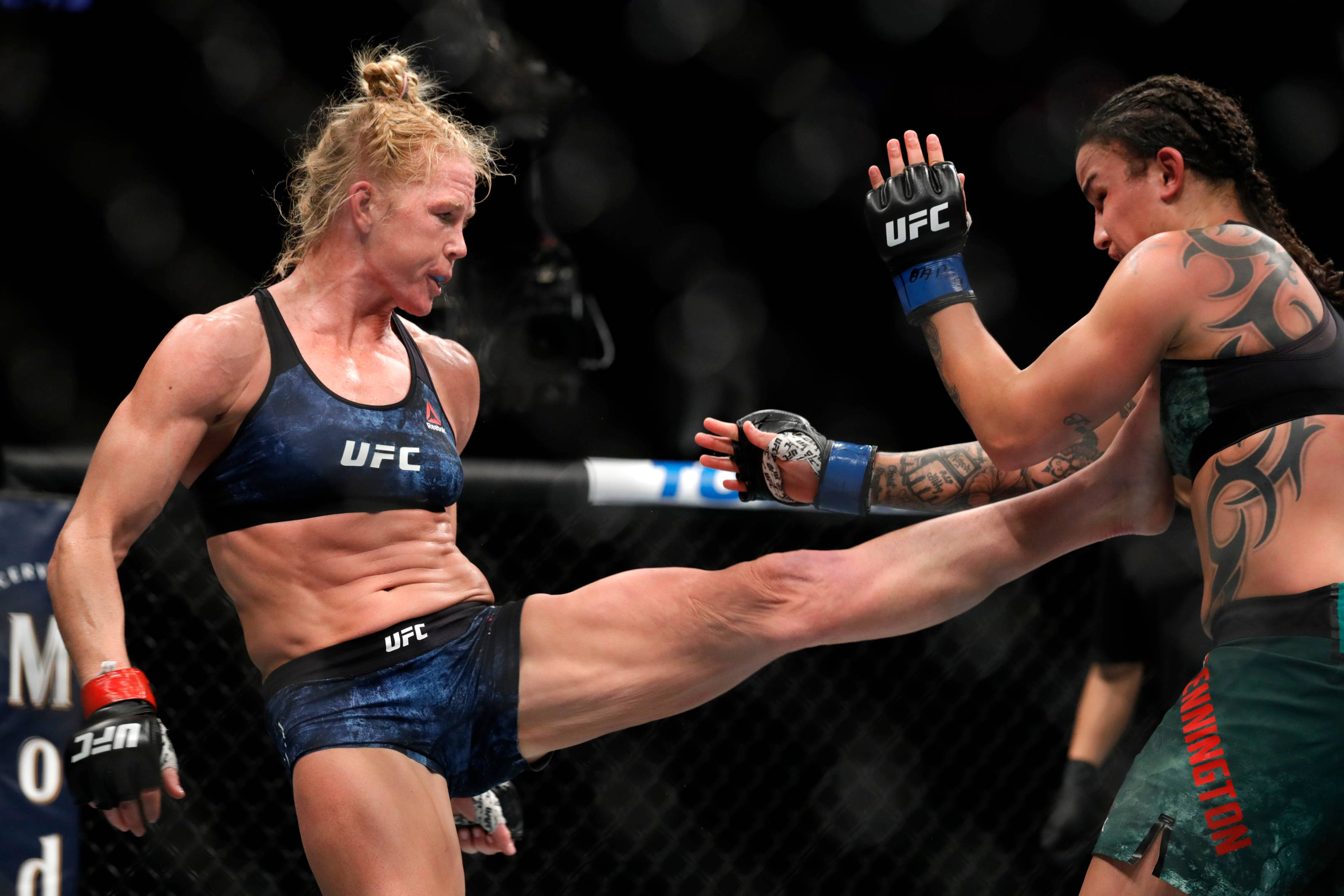 MMA 桌布 壁紙 — 兩名女性MMA拳手在籠中交鋒，一人使出高踢，另一人用拳套和可見的刺青阻擋。
