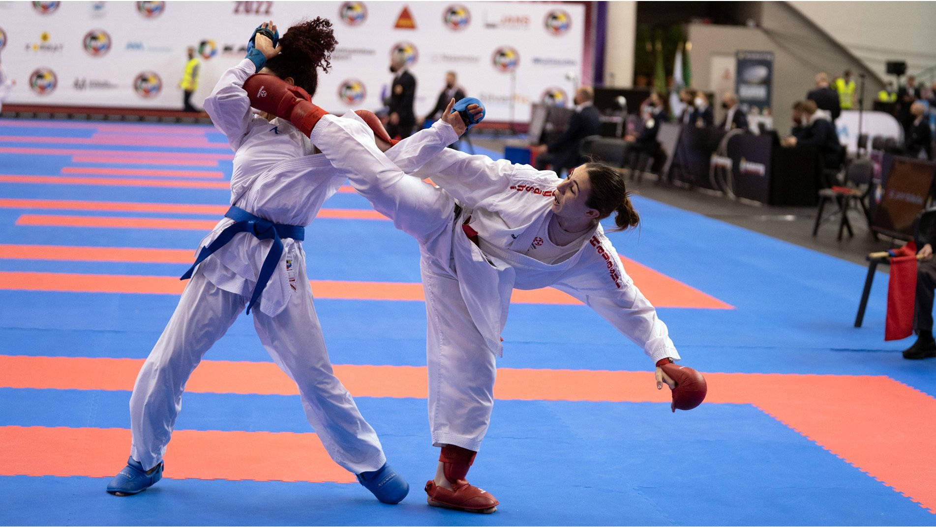 運動 桌布 壁紙 — 兩名女性跆拳道選手在藍色和橙色的比賽墊上，在明亮的場館燈光下，於踢腿中途交鋒。