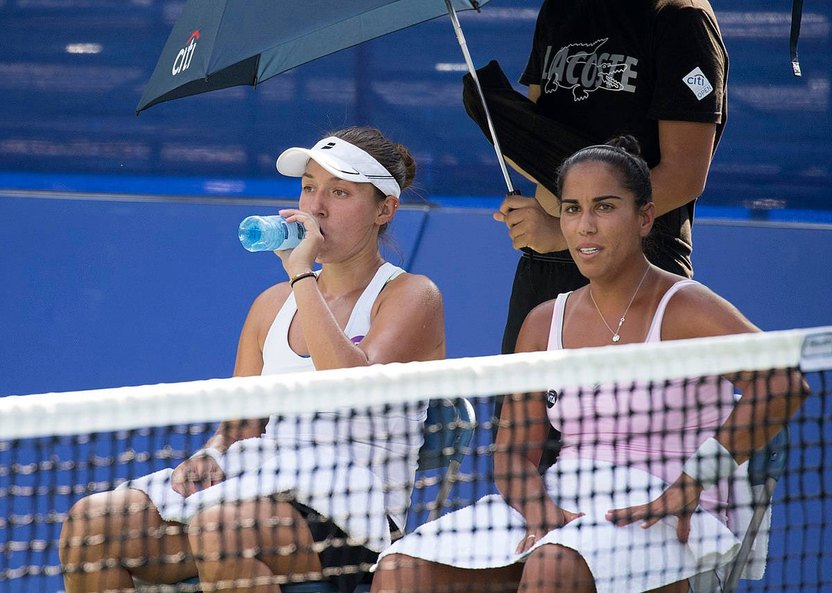 Tennis 桌布 壁紙 — 兩名女網球選手在球網後的椅子上休息，一人正在傘下喝水，另一人則向前看。