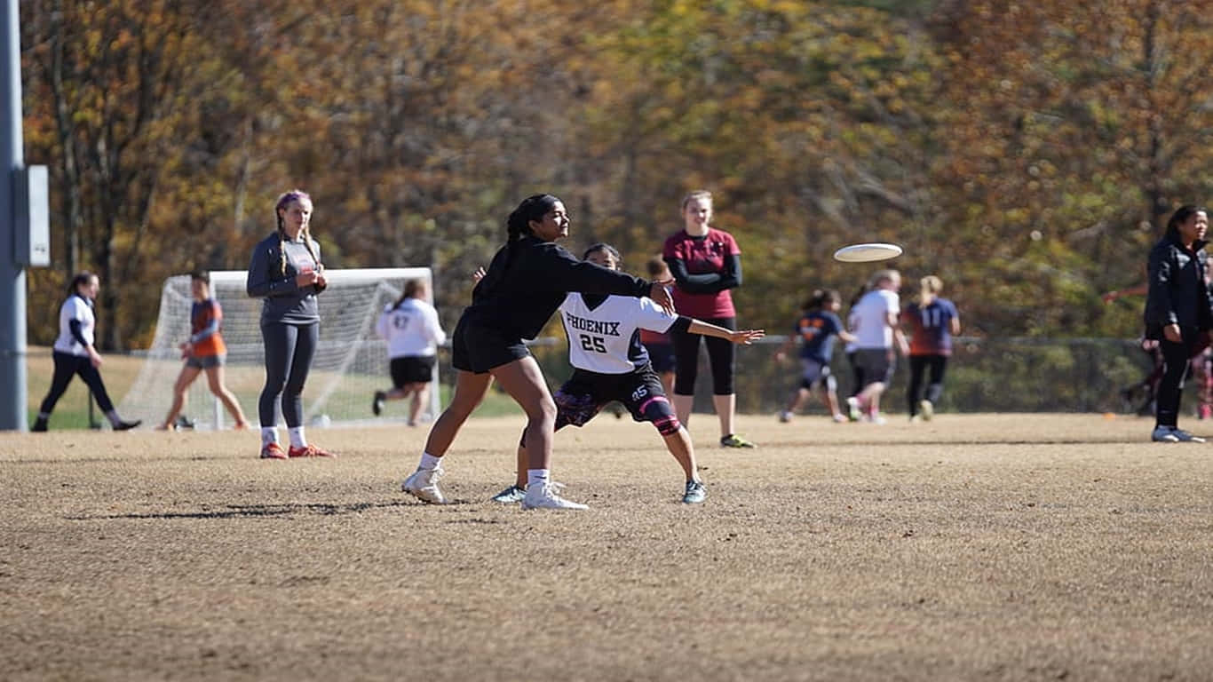 1366x768 Ultimate Frisbee 桌布 壁紙 — 兩名女運動員在陽光明媚的秋日場地上激烈爭奪一個飛行的飛盤，背景有隊友和樹木。