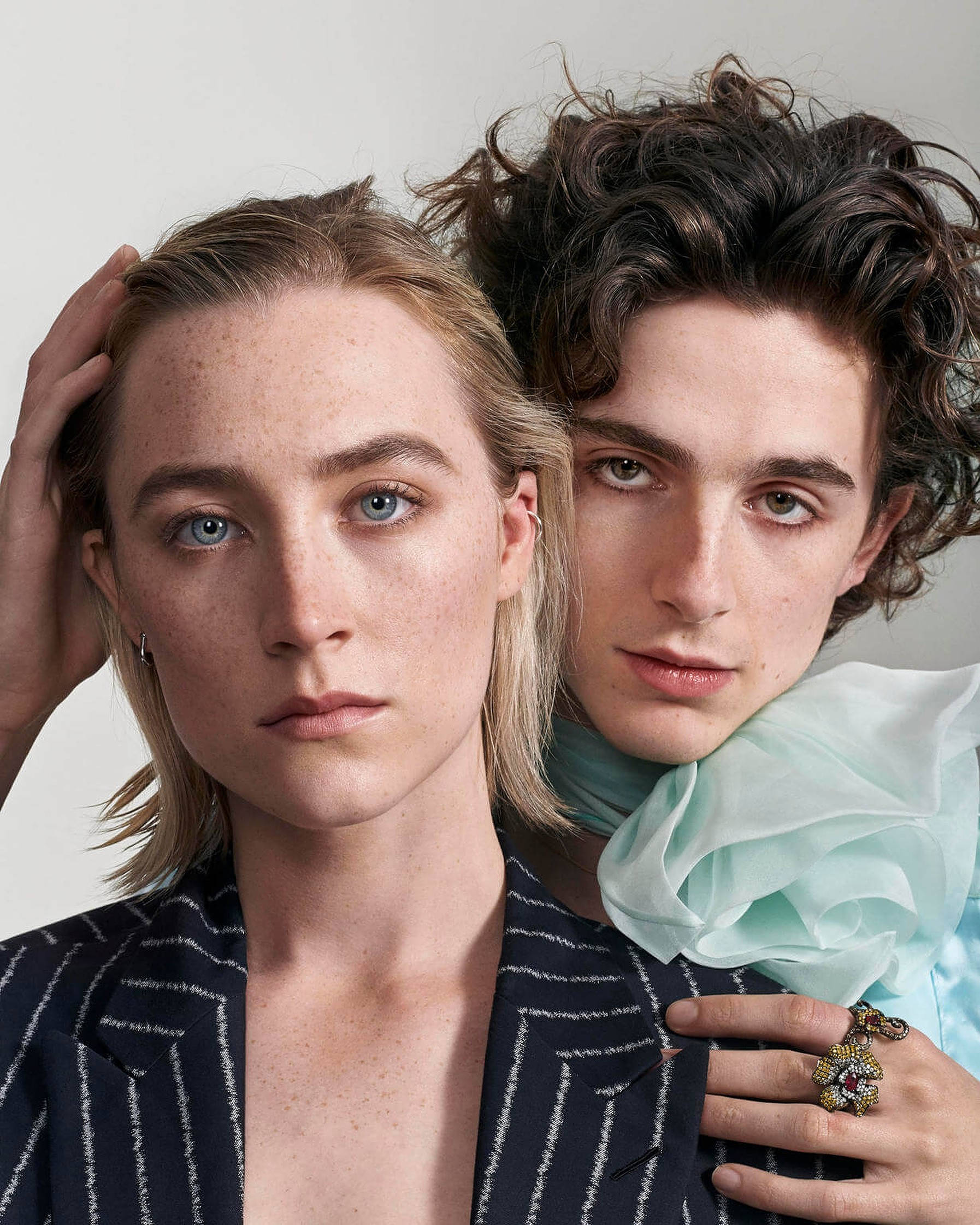 Saoirse Ronan 桌布 壁紙 — 兩名年輕成年人，臉上帶有雀斑和富有表情的眼睛，在純色背景前親密擺姿勢，一隻手放在另一人的頭上。