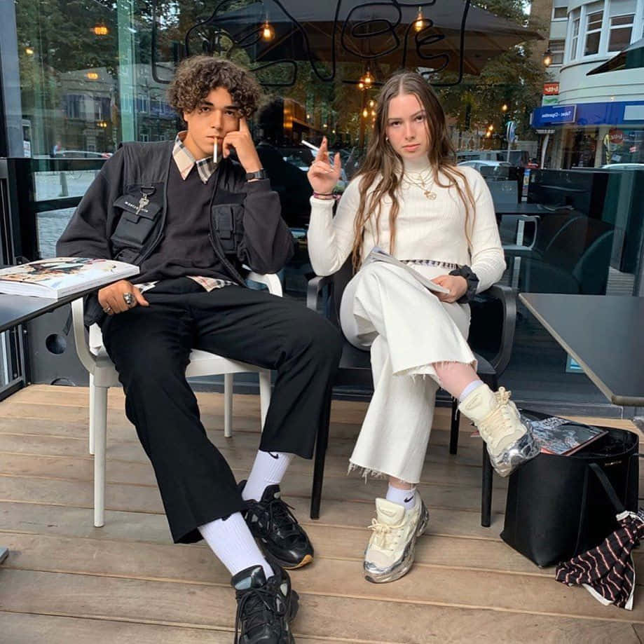 Aesthetic Couple 桌布 壁紙 — 兩名年輕成年人悠閒地坐在咖啡館的木質露台上，一人身穿黑色街頭服飾吸煙，另一人身穿白色家居服手持香煙。