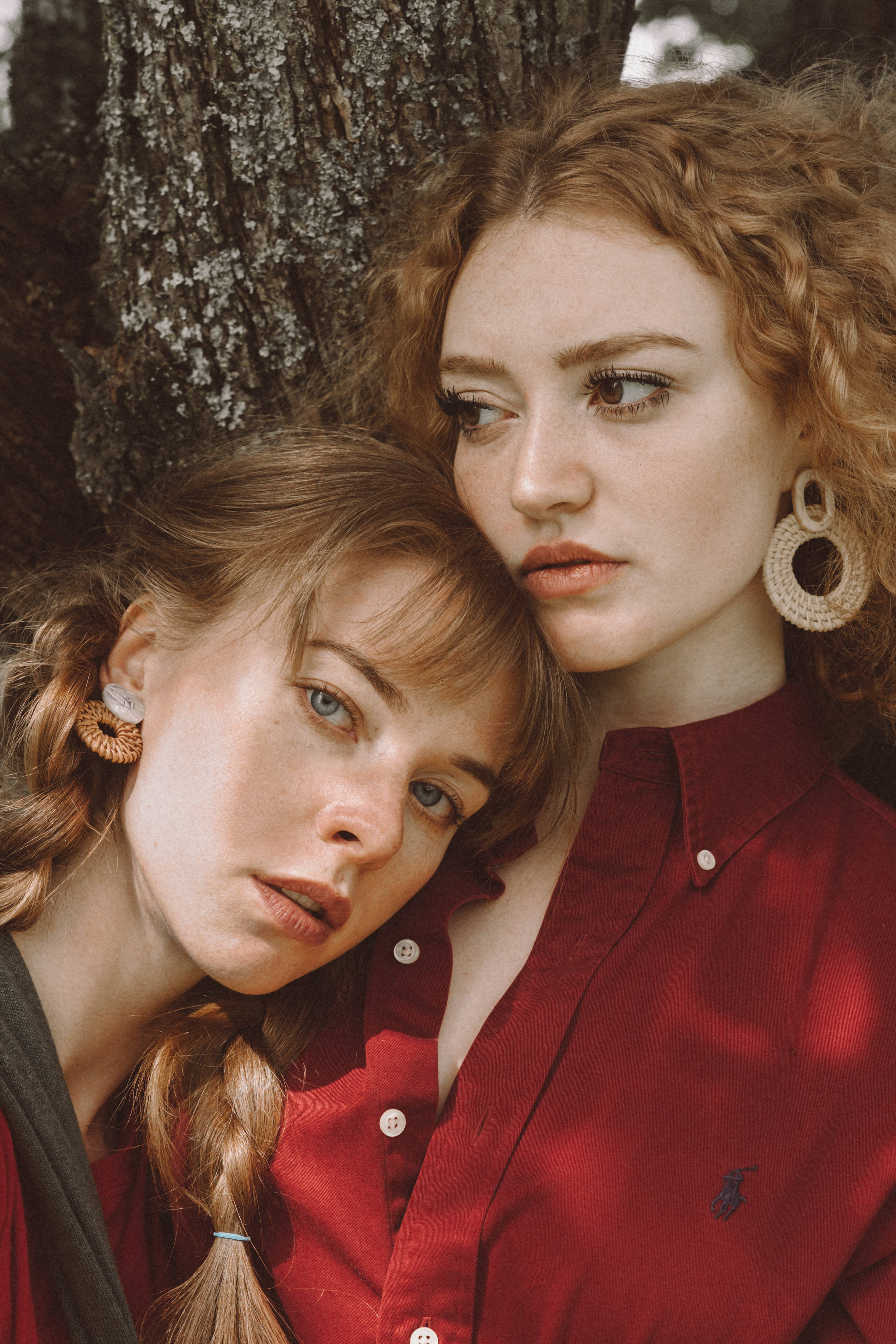 lesbians 桌布 壁紙 — 兩名年輕捲髮女性靠著樹幹，身穿紅色襯衫和編織耳環，在柔和自然光下。
