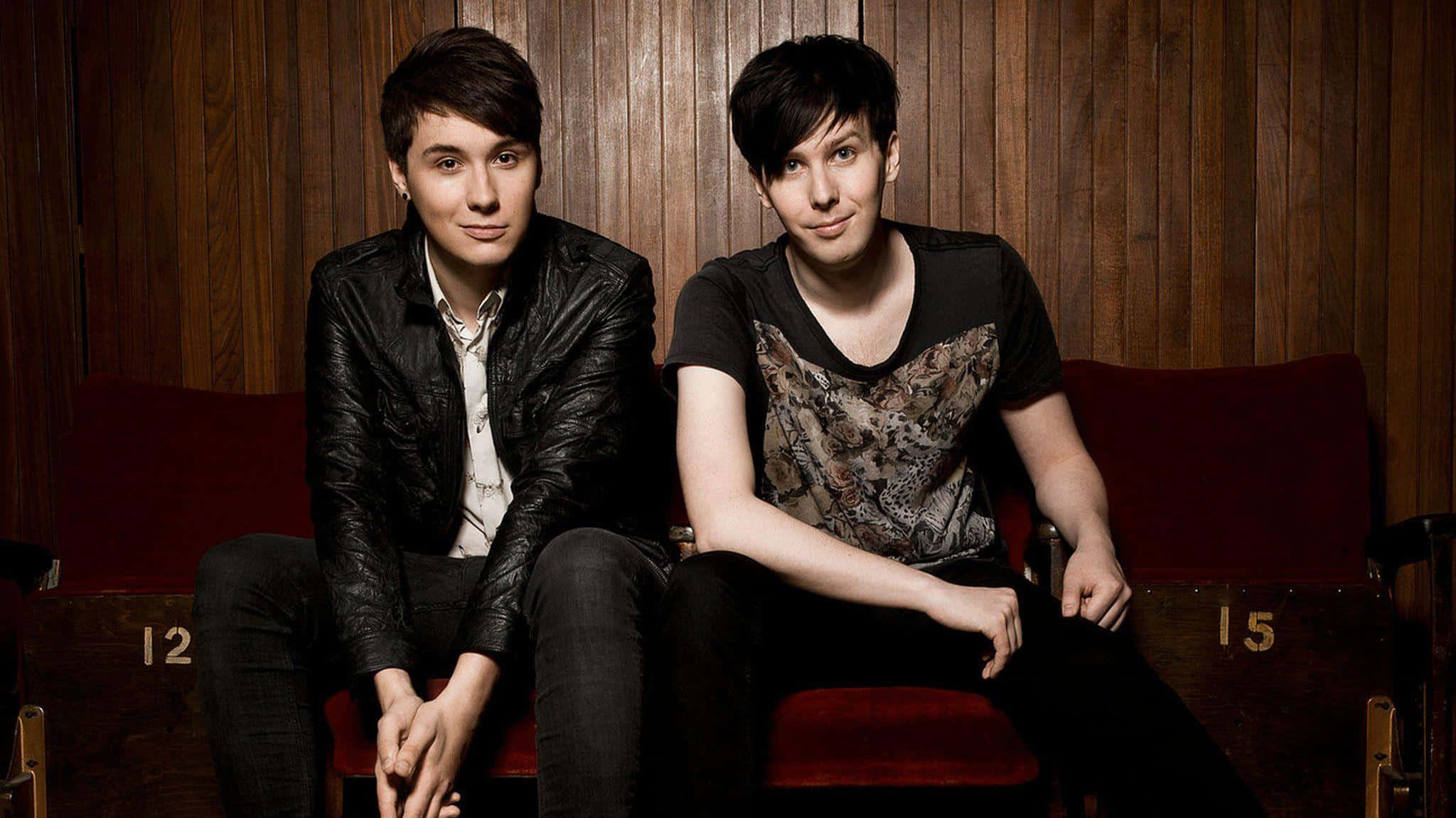 dan and phil 桌布 壁紙 — 兩名年輕男子並排坐在紅色劇院座位上，背景是溫暖的木鑲板牆。