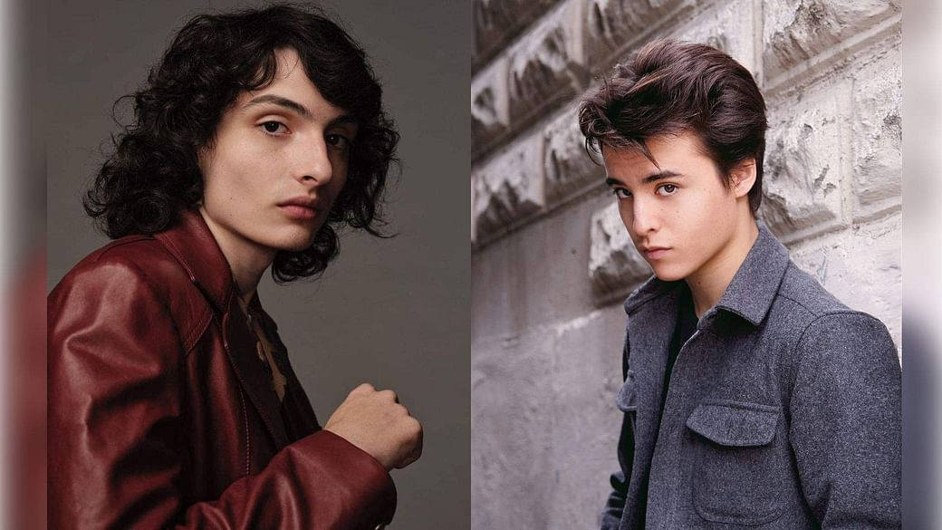 finn wolfhard 桌布 壁紙 — 兩名年輕男子擺出強烈眼神的姿勢，一人身穿紅色皮夾克，背景為灰色，另一人身穿灰色外套，靠著石牆。