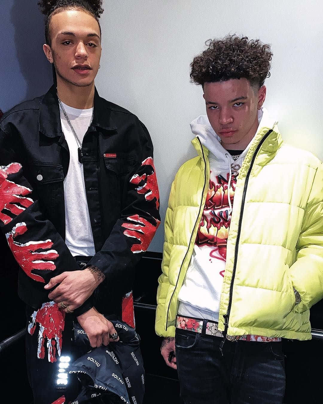 Lil Mosey 桌布 壁紙 — 兩名年輕男子穿著大膽街頭服飾，在素色牆前擺姿勢，一人穿黑色外套帶紅色手印，另一人穿螢光黃羽絨外套。
